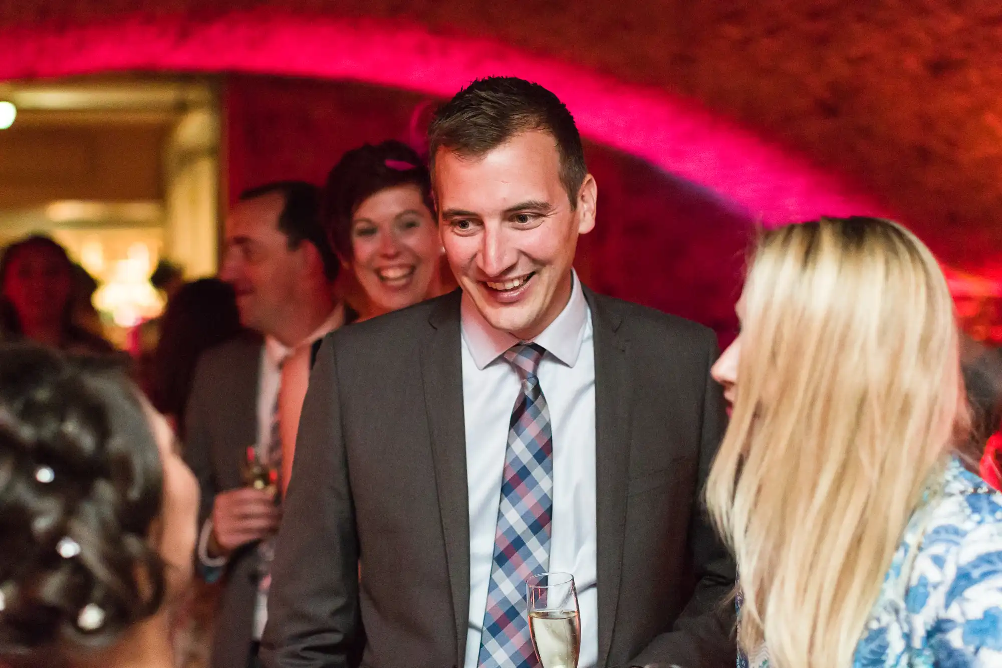 A man in a suit and checked tie smiles whilst holding a glass of champagne, talking with two women at a lively indoor event. Warm pink and red lighting creates a festive atmosphere, and several people are visible chatting in the softly focused background.