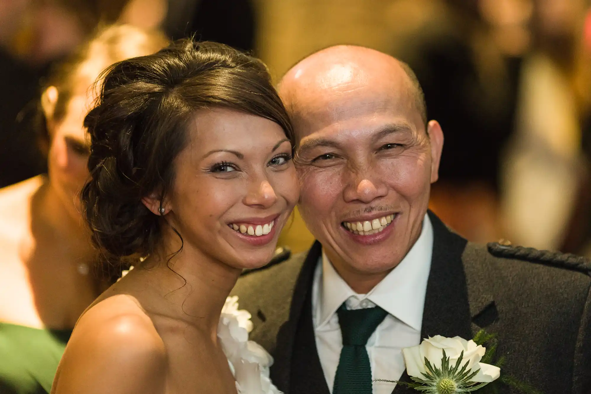 A smiling woman in a white dress and a man in a dark suit with a buttonhole stand close together, posing for a photo at what appears to be a formal event or wedding. Both look directly at the camera with warm, happy expressions.