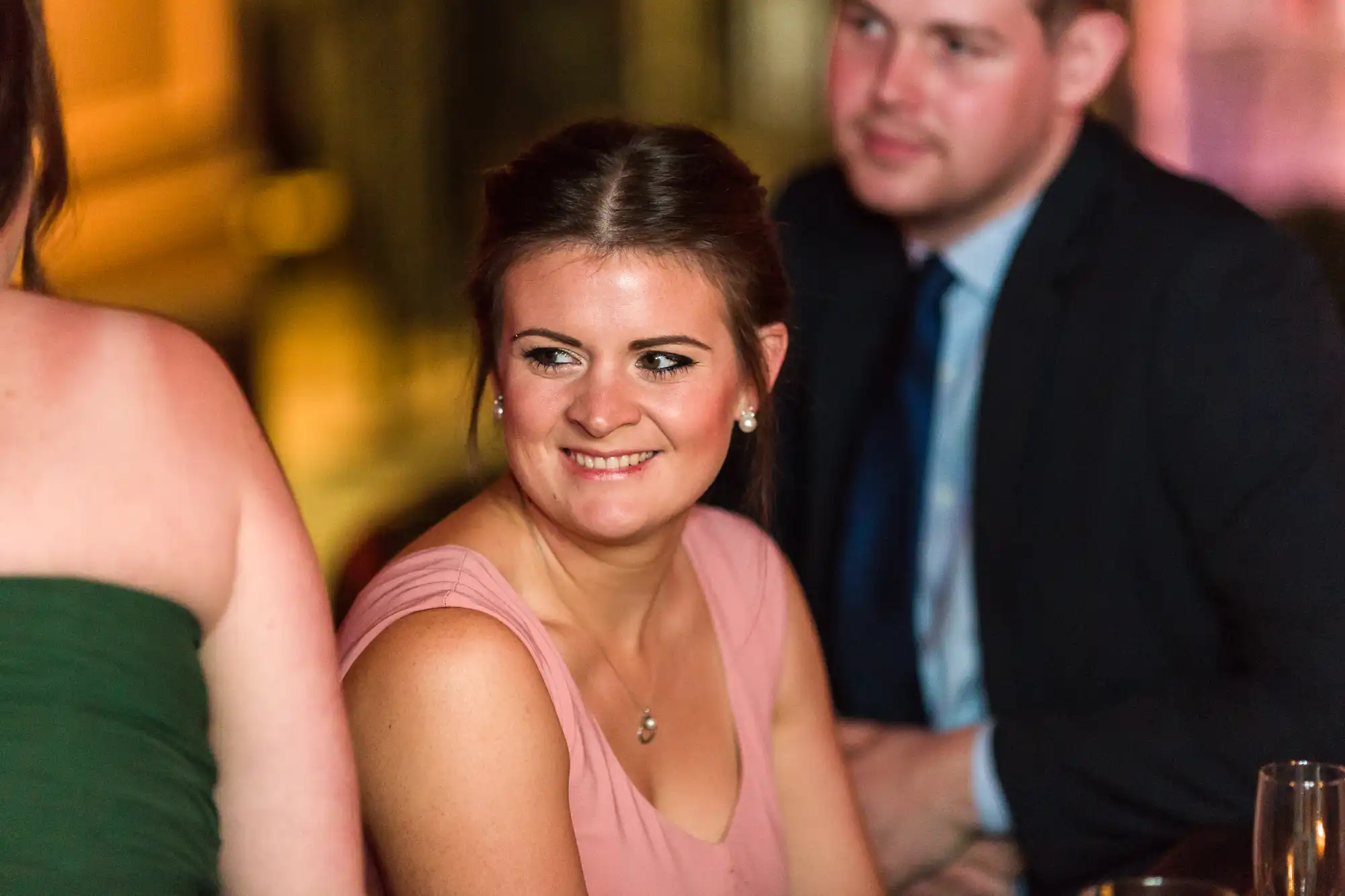 A woman in a pink sleeveless dress smiles whilst sitting at a table, looking at someone in a green dress. A man in a dark suit and tie sits blurred in the background. The setting appears warmly lit, likely at a formal or social event.