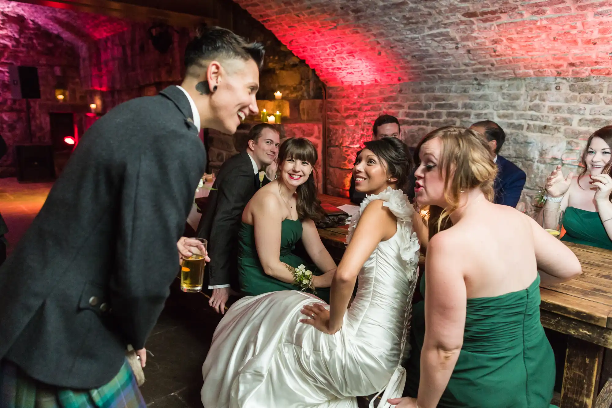 A bride in a white dress sits with bridesmaids in green dresses at a rustic, brick-vaulted venue. They smile and chat with a man in a kilt holding a drink. Warm lighting and stone walls create a cosy, festive atmosphere.