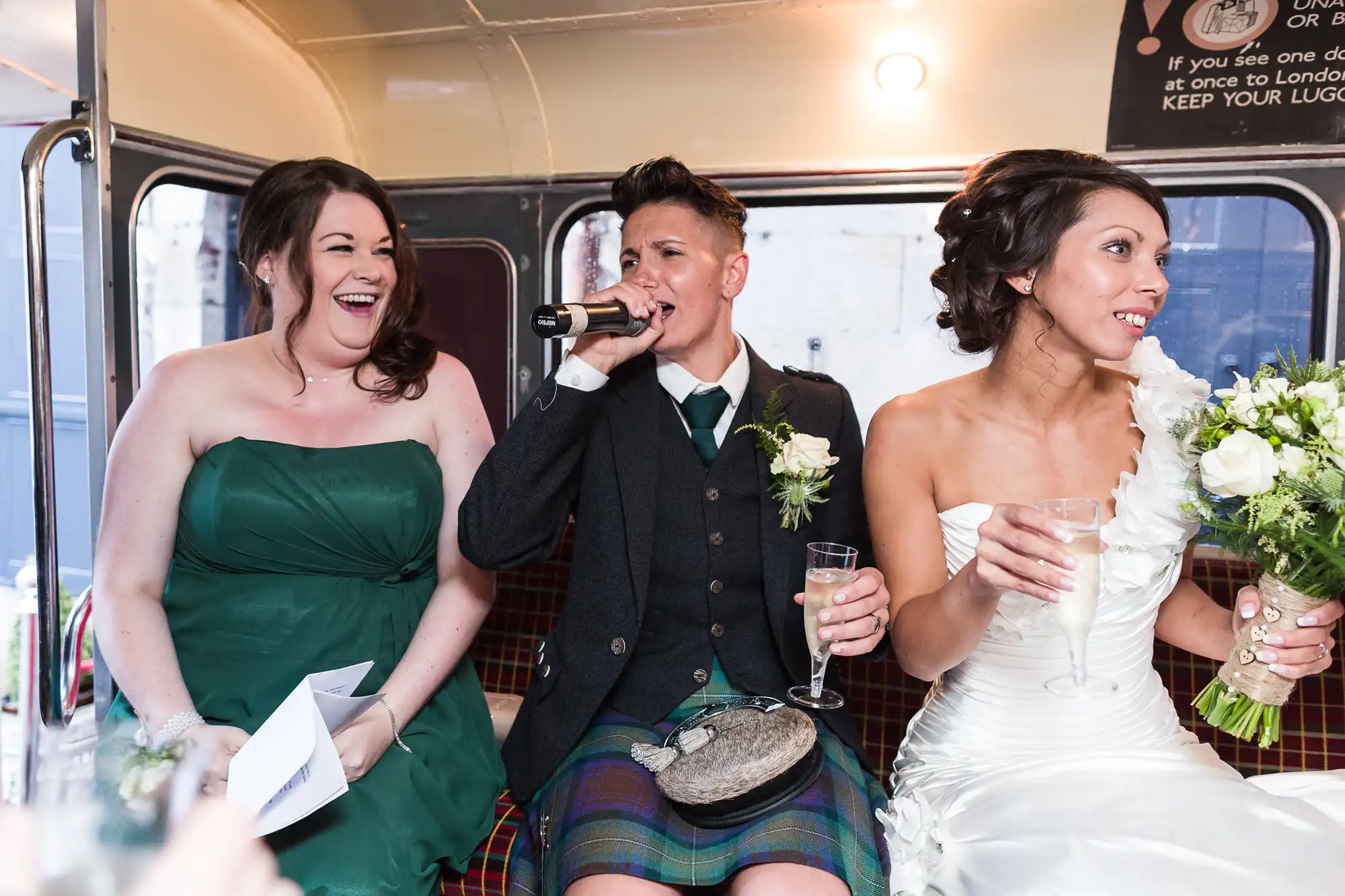 Three people sit on a bus seat dressed for a wedding. The person in the centre, wearing a suit and kilt, speaks into a microphone. The person on the left in a green dress laughs, whilst the person on the right in a white wedding dress holds flowers and a drink.