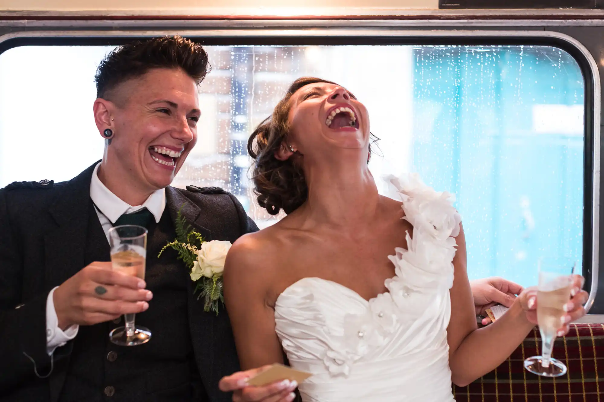 Two people dressed in formal wedding attire sit laughing joyfully on a bus. One wears a dark suit with a buttonhole and holds champagne; the other, in a white dress with floral detail, tilts their head back in laughter, also holding a glass. Rain dots the window behind them.