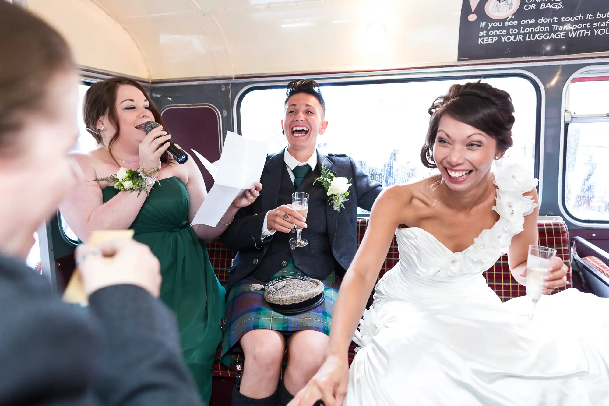 A bride in a white dress laughs joyfully whilst holding hands with a groom in a kilt and dinner jacket. Next to them, a bridesmaid with a microphone reads from a piece of paper, smiling. They’re sitting on a bus with red tartan seats, sharing a lively, celebratory moment.