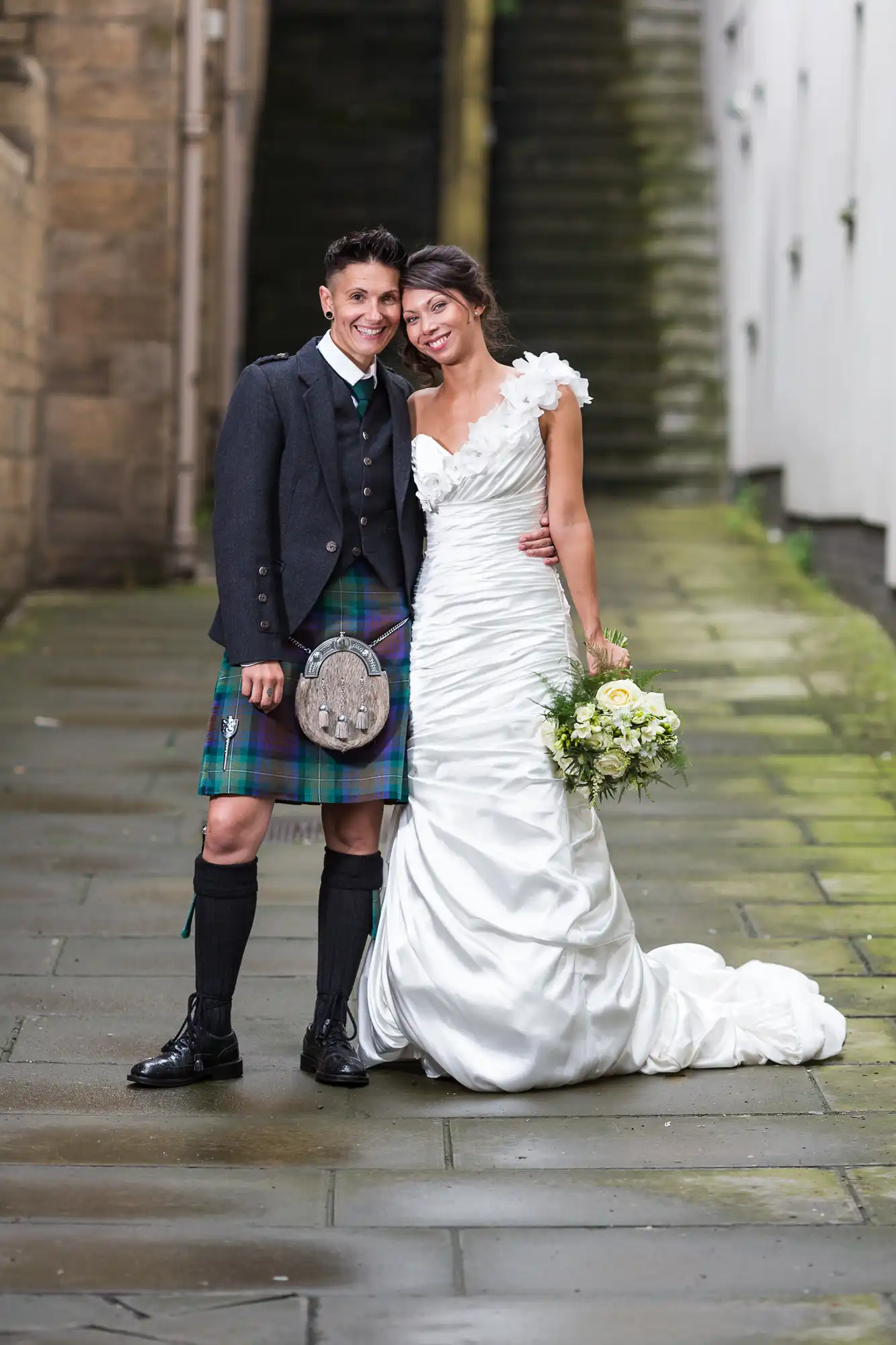 A smiling couple pose outdoors on a stone path. One person wears a dark jacket, plaid kilt, and sporran; the other wears a white wedding dress and holds a bouquet of white and yellow flowers. They stand close together, looking happy. Stone steps are in the background.