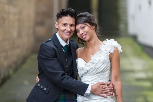 The Rowantree wedding photographer photo of two brides posing closely and smile at the camera in an outdoor setting. One wears a dark jacket and tie with a kilt, and the other wears a white, one-shoulder wedding dress with floral details. They appear happy and affectionate on their wedding day.
