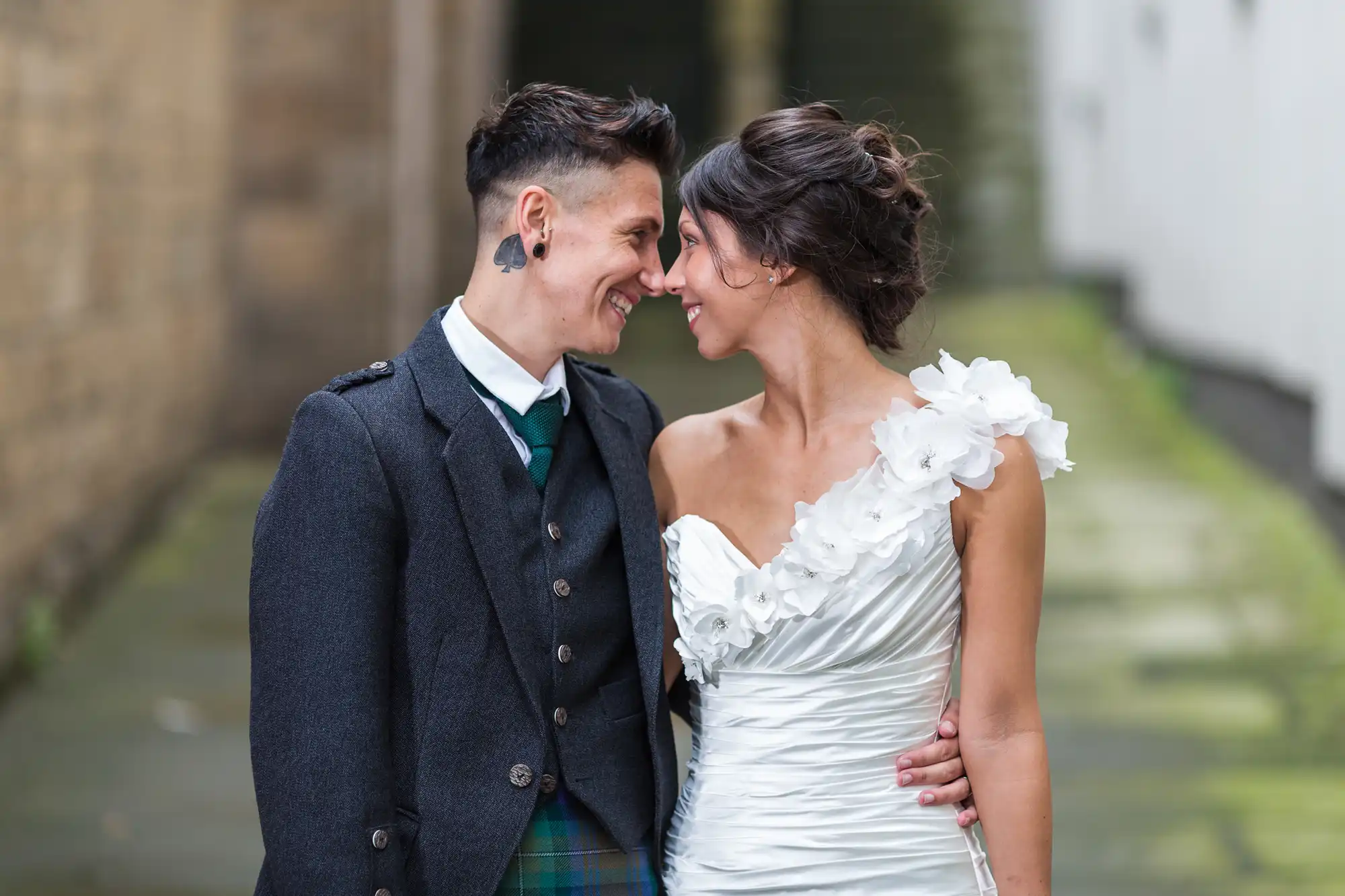 Two people in wedding attire stand close together, smiling and touching foreheads. One wears a dark suit with a green tie and tartan kilt, the other a white, one-shoulder dress with floral details. They appear joyful in an outdoor, urban alleyway.