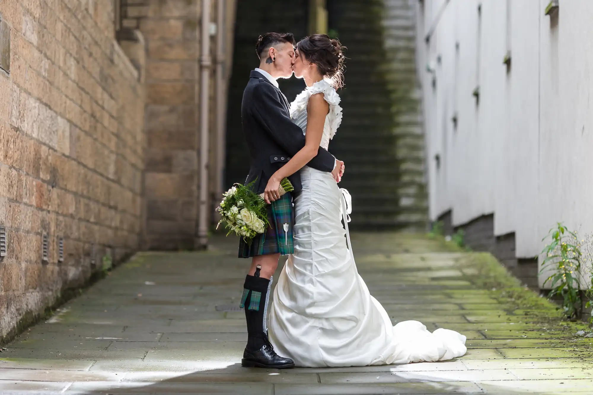 A couple share a kiss in a narrow, sunlit alleyway. The person on the left wears a black jacket and tartan kilt, holding a bouquet. The person on the right wears a white, ruched wedding dress with a train. They embrace, surrounded by stone walls and steps in the background.