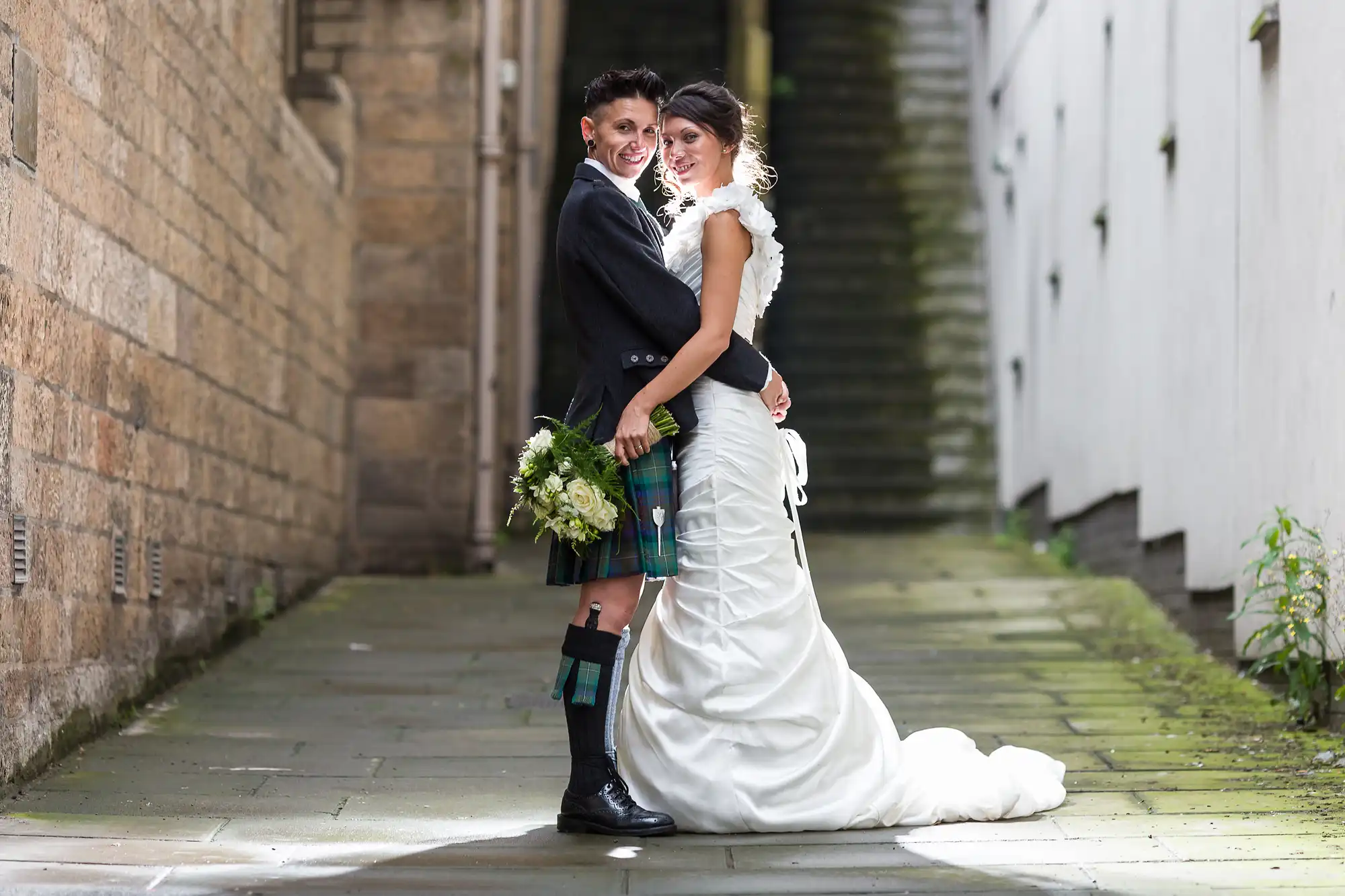 A smiling couple stands in a narrow alleyway. One wears a black jacket, kilt, and boots while holding a bouquet; the other wears a white wedding dress with a long train. Sunlight highlights them, with stone and white walls on either side and steps behind.