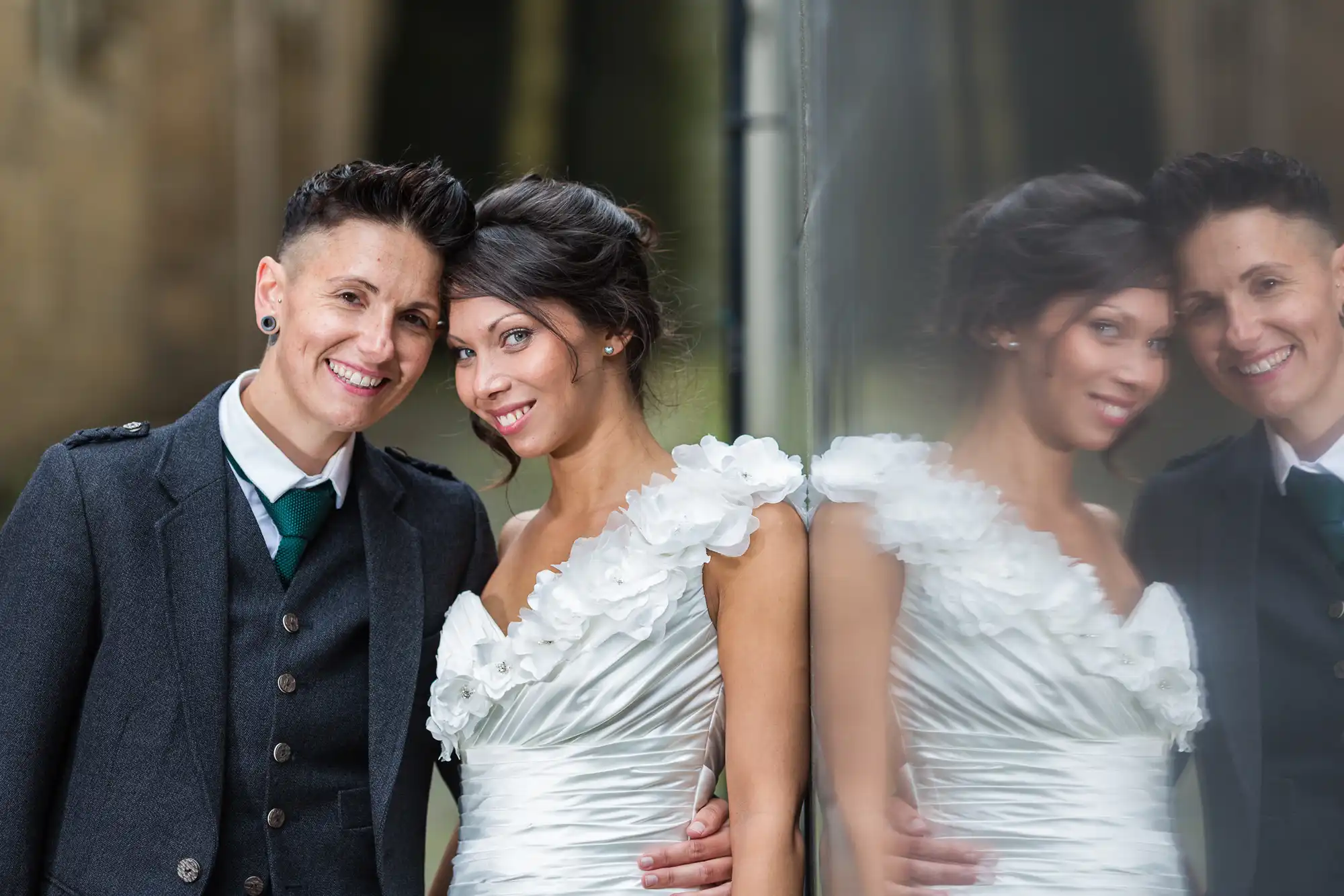 Two women pose closely, smiling, in wedding attire. One wears a dark suit and tie, the other a white dress with flower details on the shoulder. They stand by a reflective surface, creating a soft reflection of their embrace, with a blurred outdoor background.