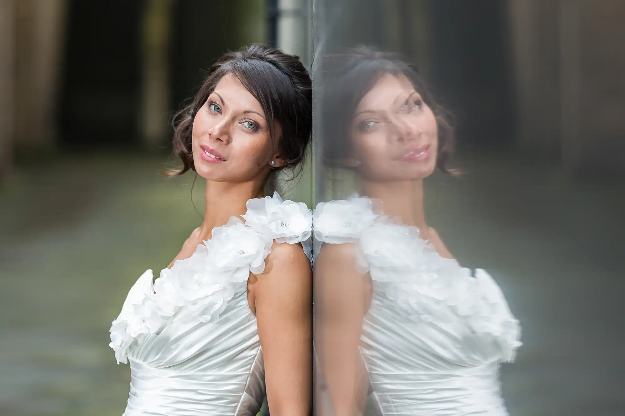 A woman in a white, floral-shouldered dress leans against a reflective glass surface, creating a mirrored image of her face and upper body. She has brown hair styled up and gazes softly at the camera against a blurred outdoor background.