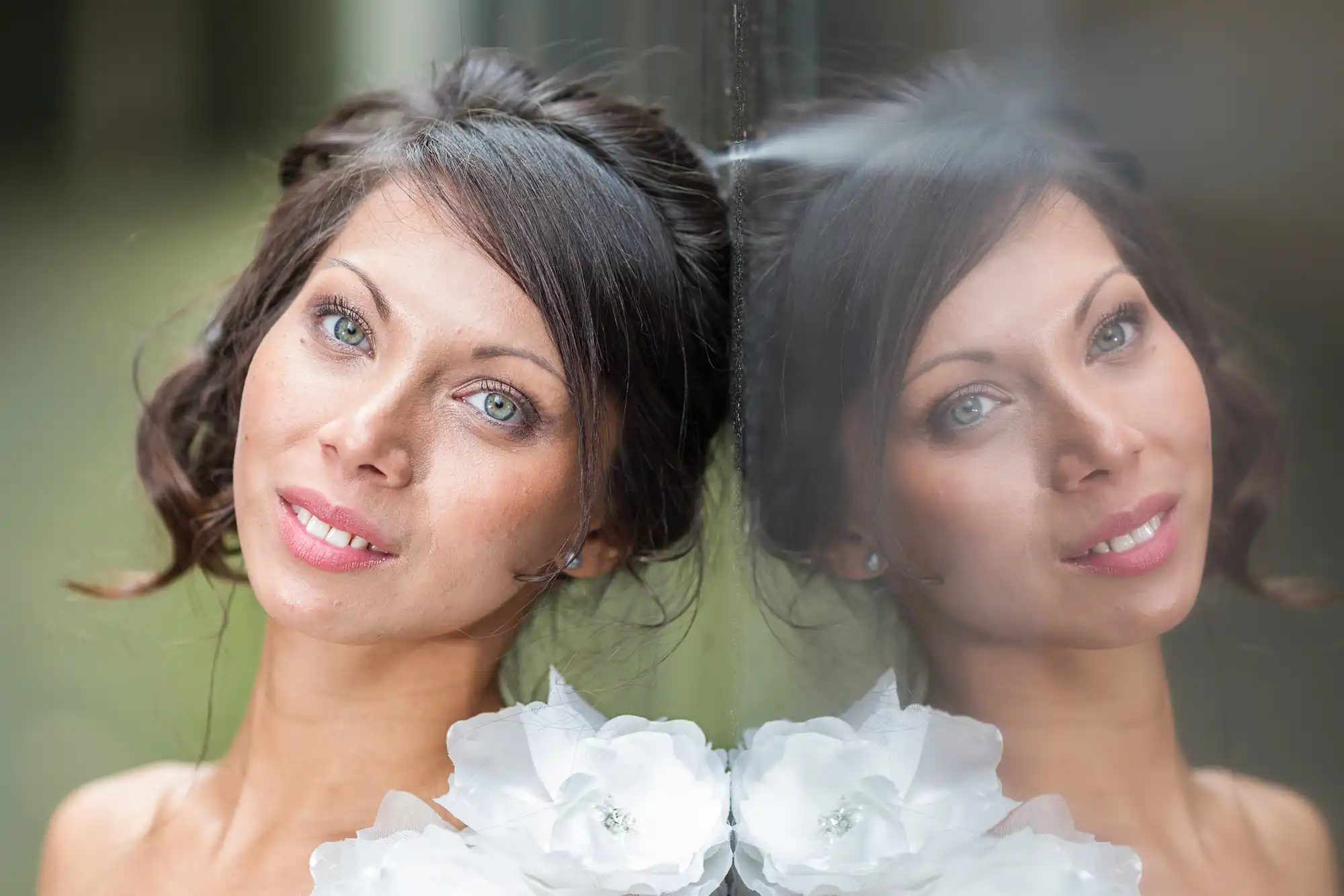 A woman with light eyes and dark hair styled up smiles gently, wearing a white dress with large floral details. She is leaning against a reflective surface, creating a clear mirror image of her face and upper body beside her.