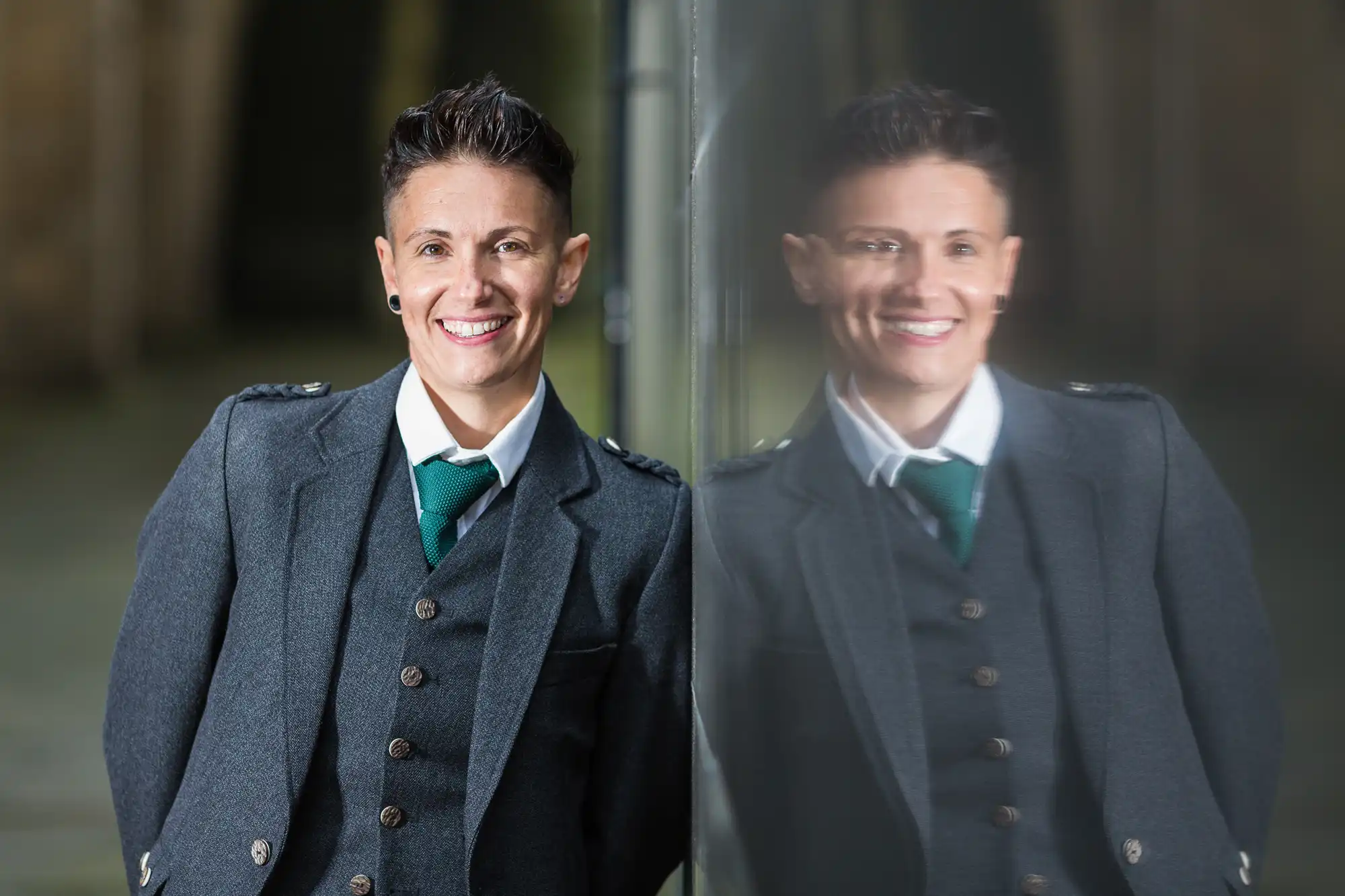 A person with short dark hair, wearing a grey suit, white shirt, and green tie, leans against a reflective glass surface. Their reflection is visible, and they are smiling at the camera. The background is softly blurred.