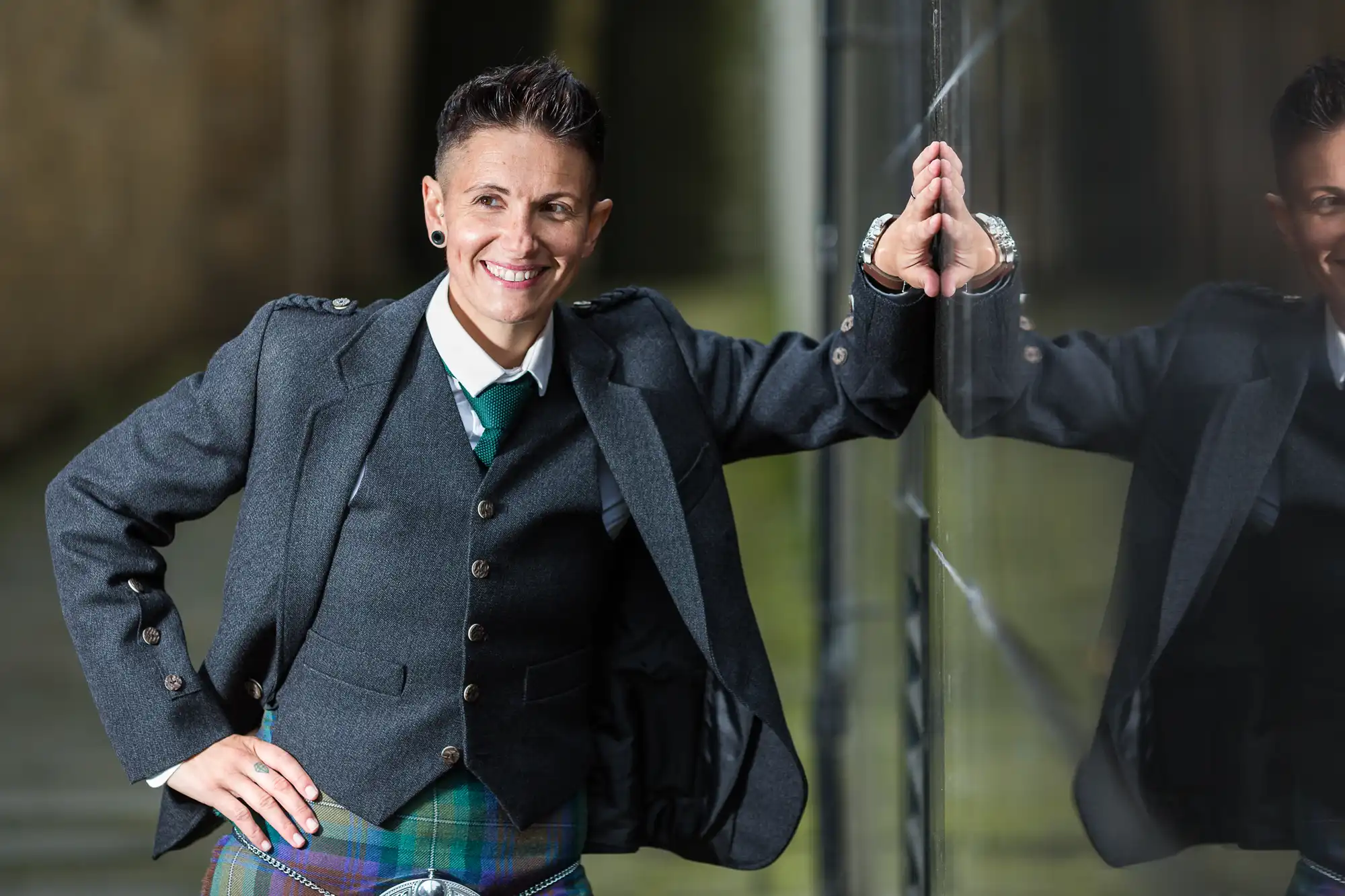 A person with short dark hair, wearing a green and blue tartan kilt, white shirt, green tie, and dark jacket, smiles whilst leaning against a reflective glass wall. Their reflection is visible, and they appear relaxed and confident outdoors.