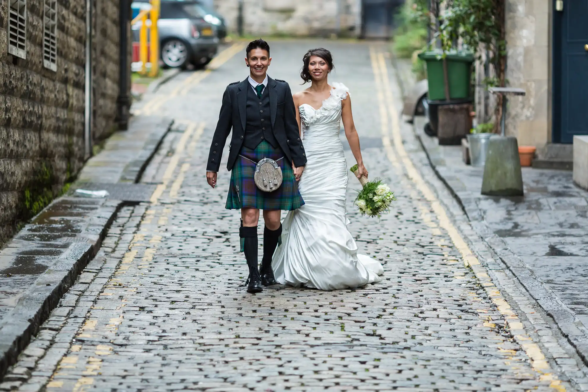 A smiling couple walk hand-in-hand on a cobblestone street. The groom wears a Scottish kilt and jacket, while the bride wears a white gown holding a bouquet. Old stone buildings and greenery line the empty street in the background.