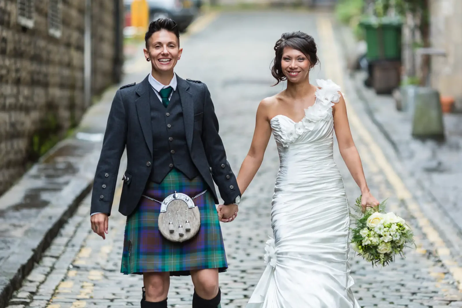 A smiling couple hold hands on a cobbled street. One person wears a grey jacket, white shirt, tie, and tartan kilt; the other wears a white, one-shouldered wedding dress and holds a bouquet of flowers. Both look happy and celebratory as they are photographed by The Rowantree wedding photographer.