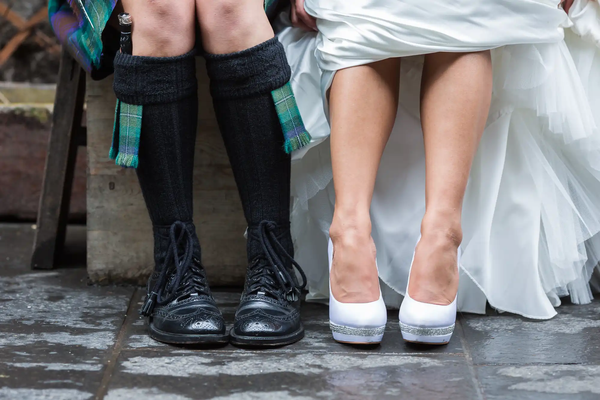Two people sit side by side, showing only their legs and feet. One wears a white wedding dress with white heels, while the other wears a kilt, knee-high socks, and black brogue shoes. The ground beneath them is stone.