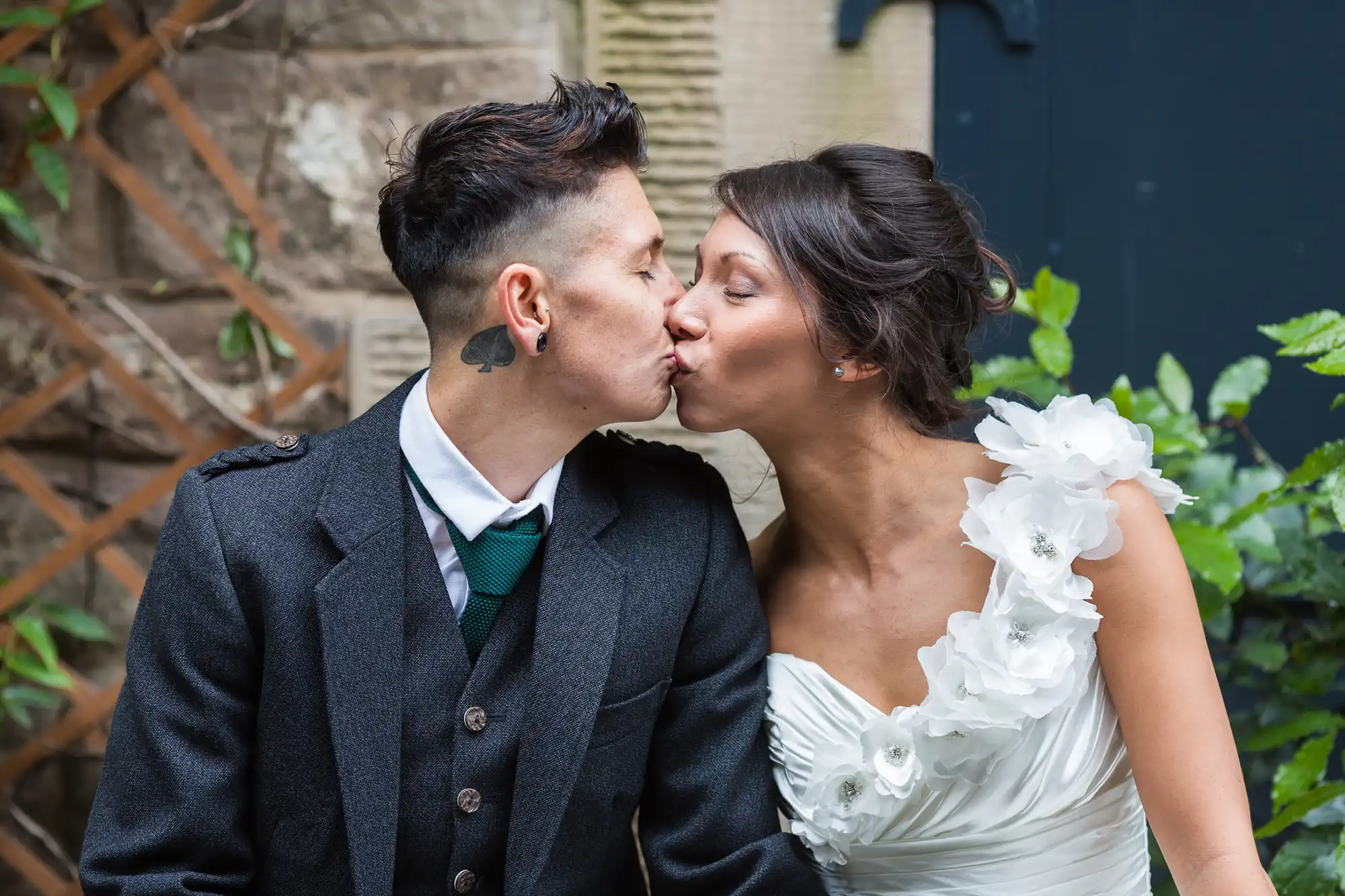 A couple in wedding attire share a kiss outdoors. One wears a dark suit, white shirt, and green tie with a heart tattoo on their neck; the other wears a white one-shoulder dress adorned with floral details. Lush greenery and stone walls are in the background.