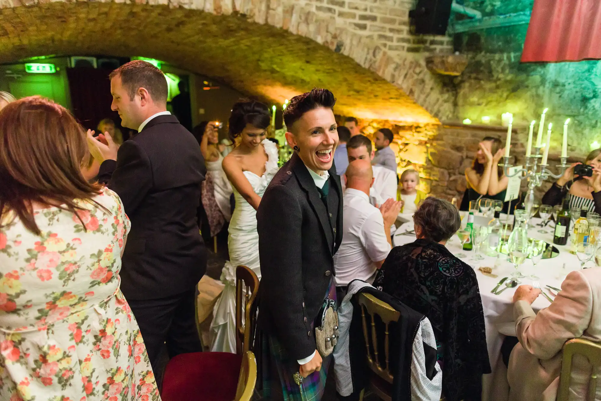 A joyful person in a black jacket and tartan kilt laughs amid a wedding reception in a stone-walled venue. Guests in formal attire sit and stand around decorated tables, whilst warm lighting creates a festive, cheerful atmosphere.