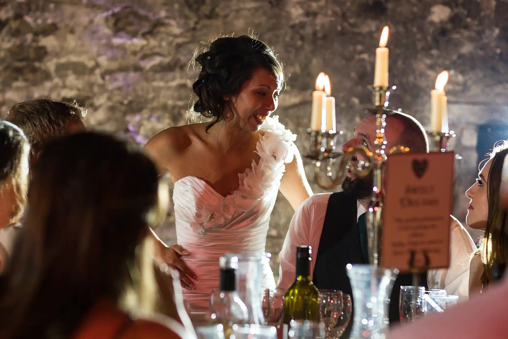A woman in a white dress with floral detail smiles and interacts with seated guests at a candlelit dinner, surrounded by glassware and bottles, in a rustic stone-walled setting. Warm light from candles creates a festive, elegant atmosphere.