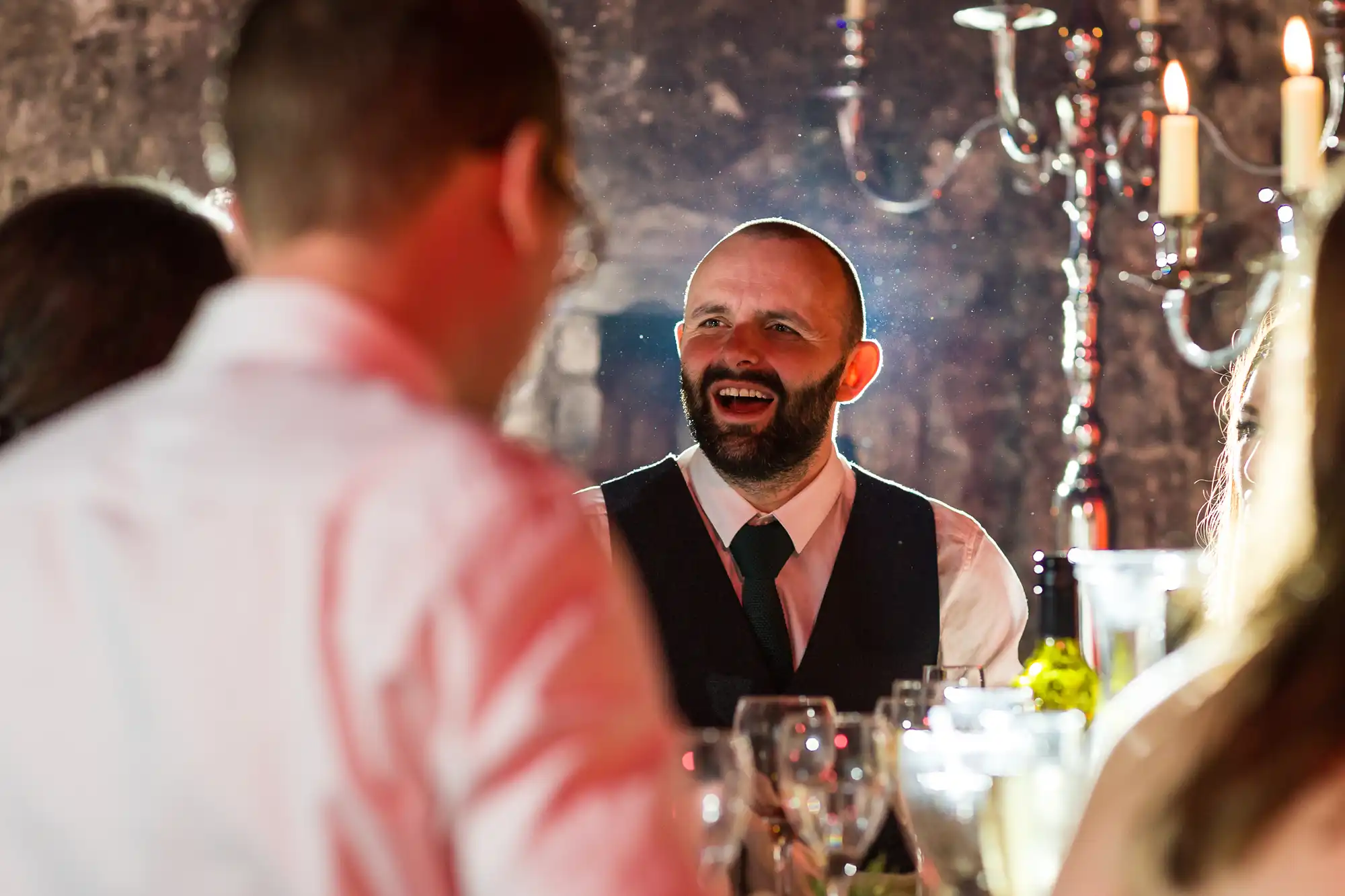 A bearded man in a dark waistcoat and tie sits at a dinner table, smiling and talking with others. The table is set with glasses and bottles. Soft lighting and a silver candelabrum in the background create a warm, festive atmosphere.