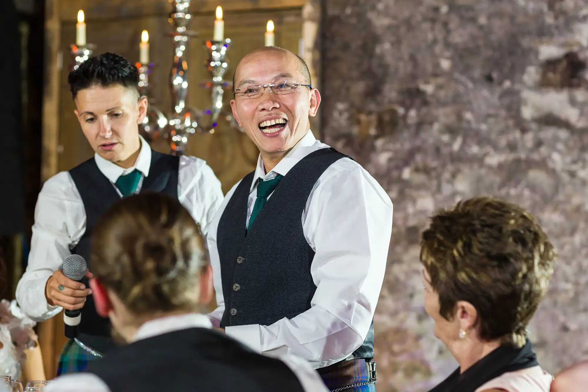 A man wearing glasses, a white shirt, green tie, and dark waistcoat stands smiling and laughing in a warmly lit room. Beside him, another person in similar attire holds a microphone. Two people, slightly blurred, sit facing them. A candelabrum glows in the background.