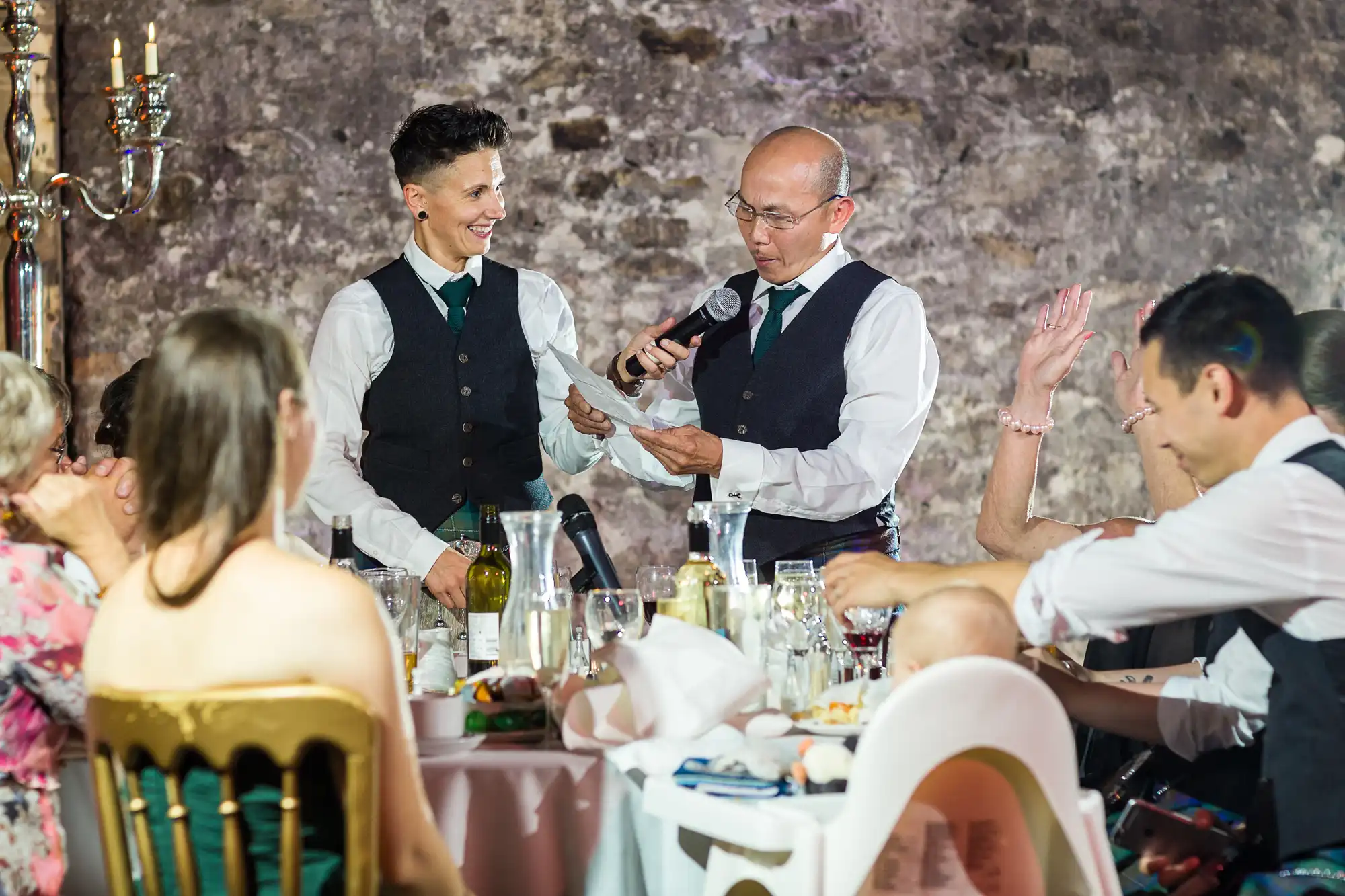 Two people in matching waistcoats and ties stand by a round table at a formal event, one holding a microphone and reading from a paper while the other smiles. Seated guests listen attentively, set against a rustic stone wall, with food and drinks on the table.
