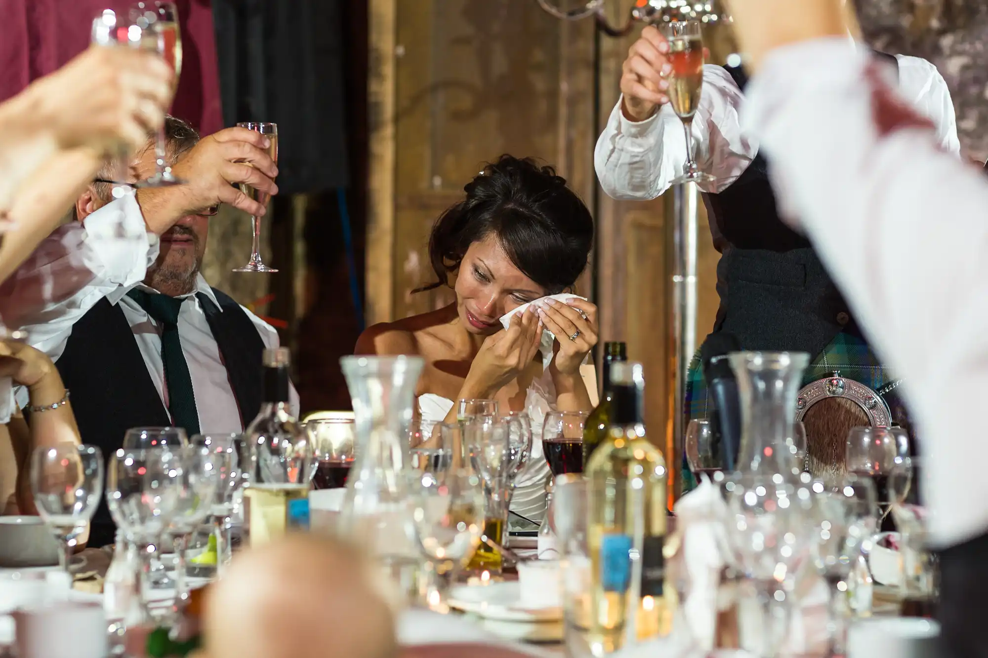 A woman in a white dress sits at a table crowded with glasses and bottles, wiping tears from her face and smiling. People around her, mostly in formal attire, raise glasses in a toast, celebrating at what appears to be a wedding reception.