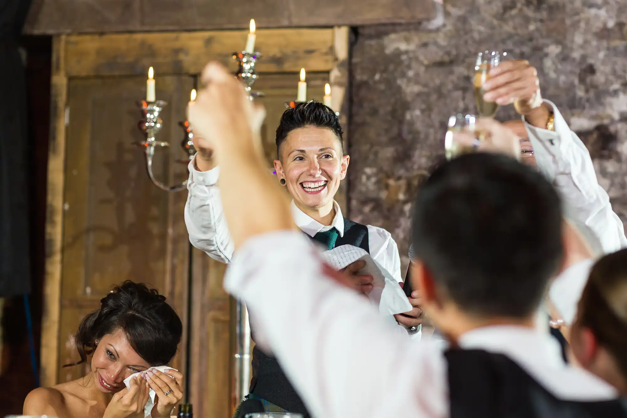 A smiling person in formal attire gives a toast, holding a piece of paper and raising a glass, while guests with raised glasses celebrate. One guest wipes away a tear, suggesting an emotional moment, and a candelabrum glows in the background.
