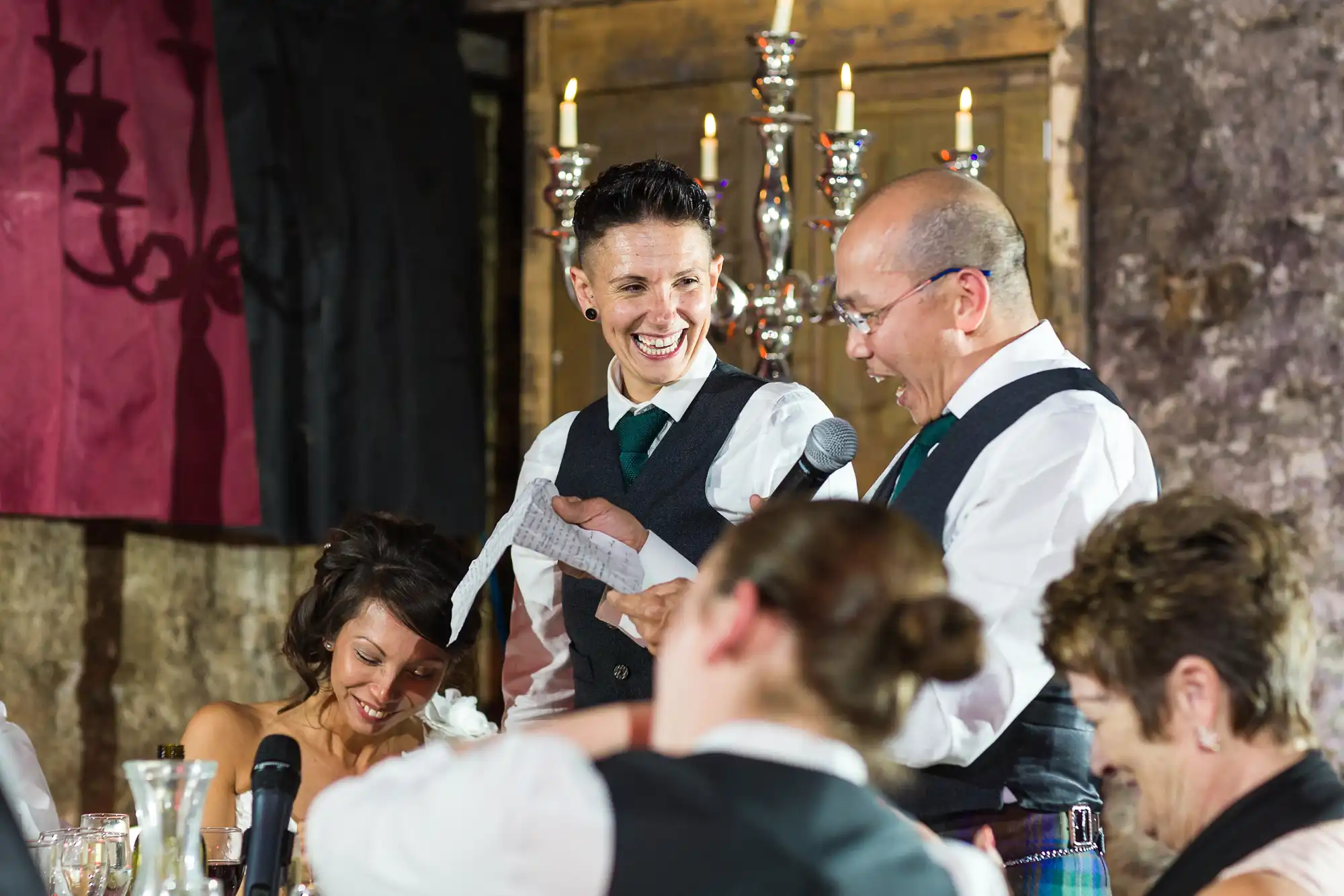 A smiling newlywed couple wearing waistcoats and ties stands at a wedding reception; one holds a microphone and paper. Guests around them laugh, and a candelabra with lit candles sits behind them against a rustic stone wall.