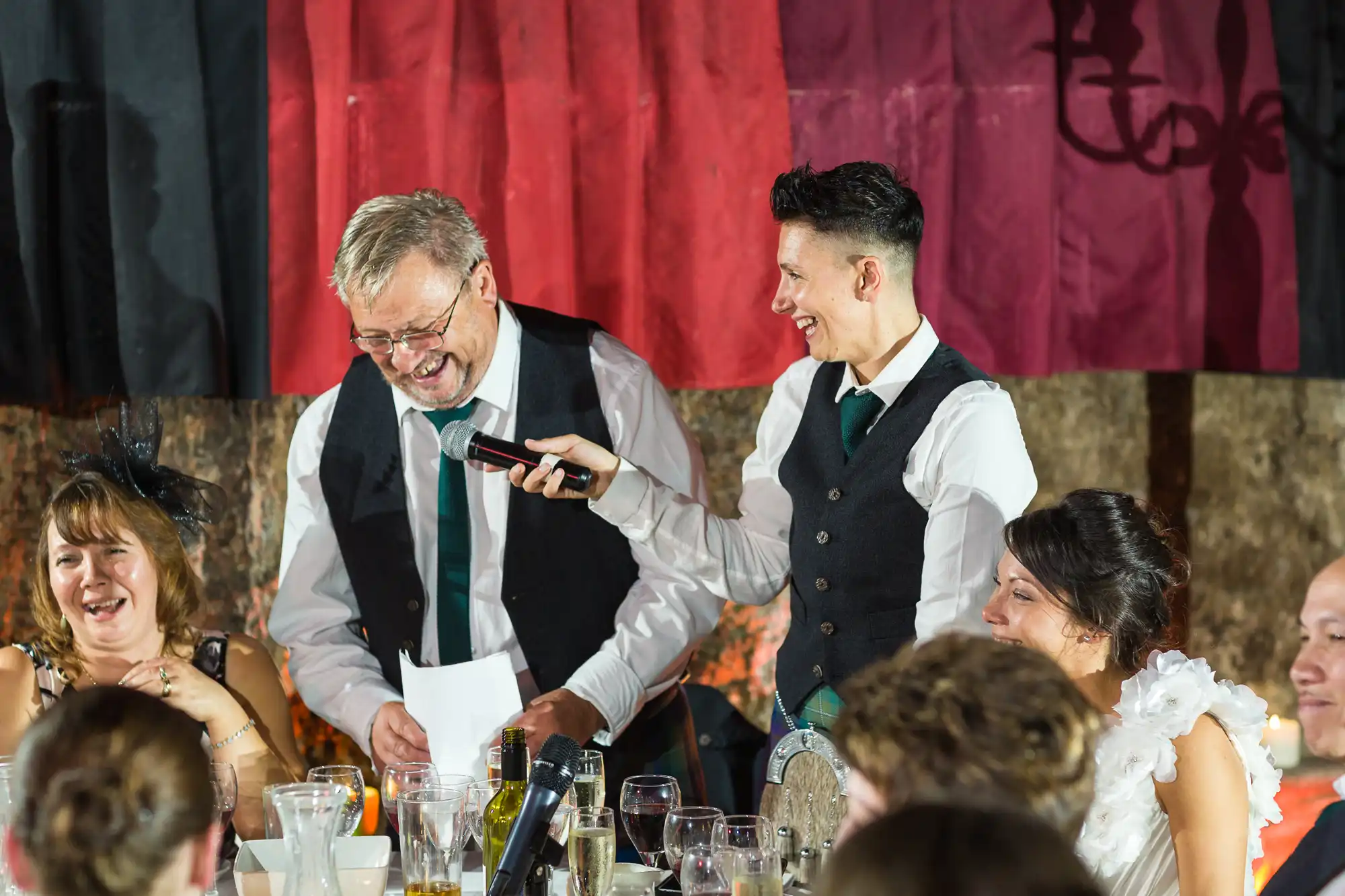 A lively group at a formal event laughs together. A person in a waistcoat and tie holds a microphone to an older man with glasses, who is smiling and holding papers. Two women nearby are also laughing, with drinks, crockery, and decorations visible. A red and black curtain hangs behind.