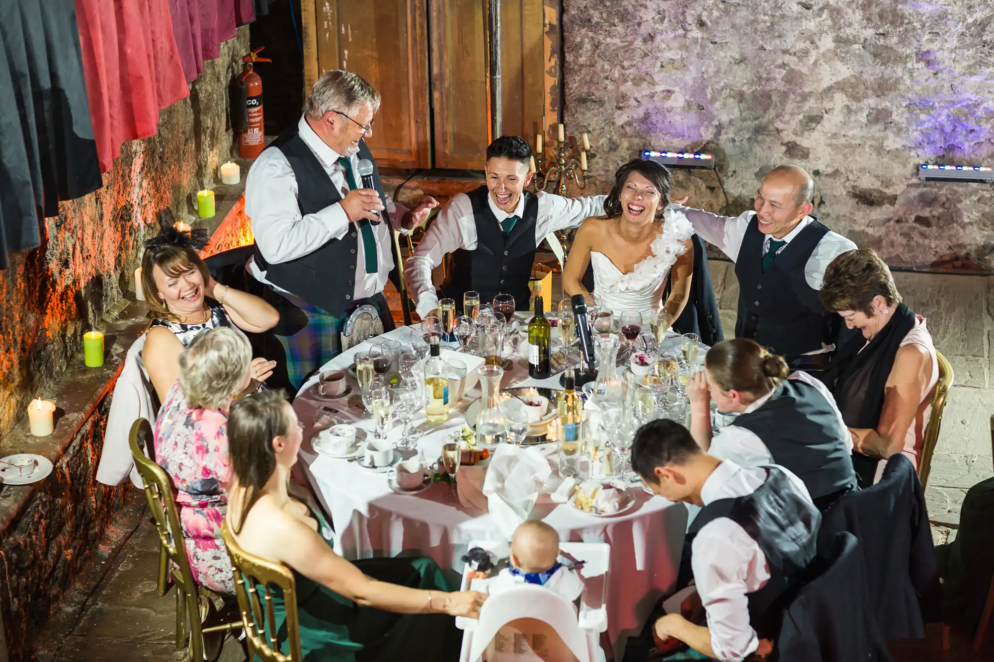 A joyful wedding reception scene shows guests laughing around a round table set with food, drinks, and flowers. The bride and groom sit side by side, smiling, while an older man stands giving a toast. Warm lighting and stone walls create a festive, intimate atmosphere.