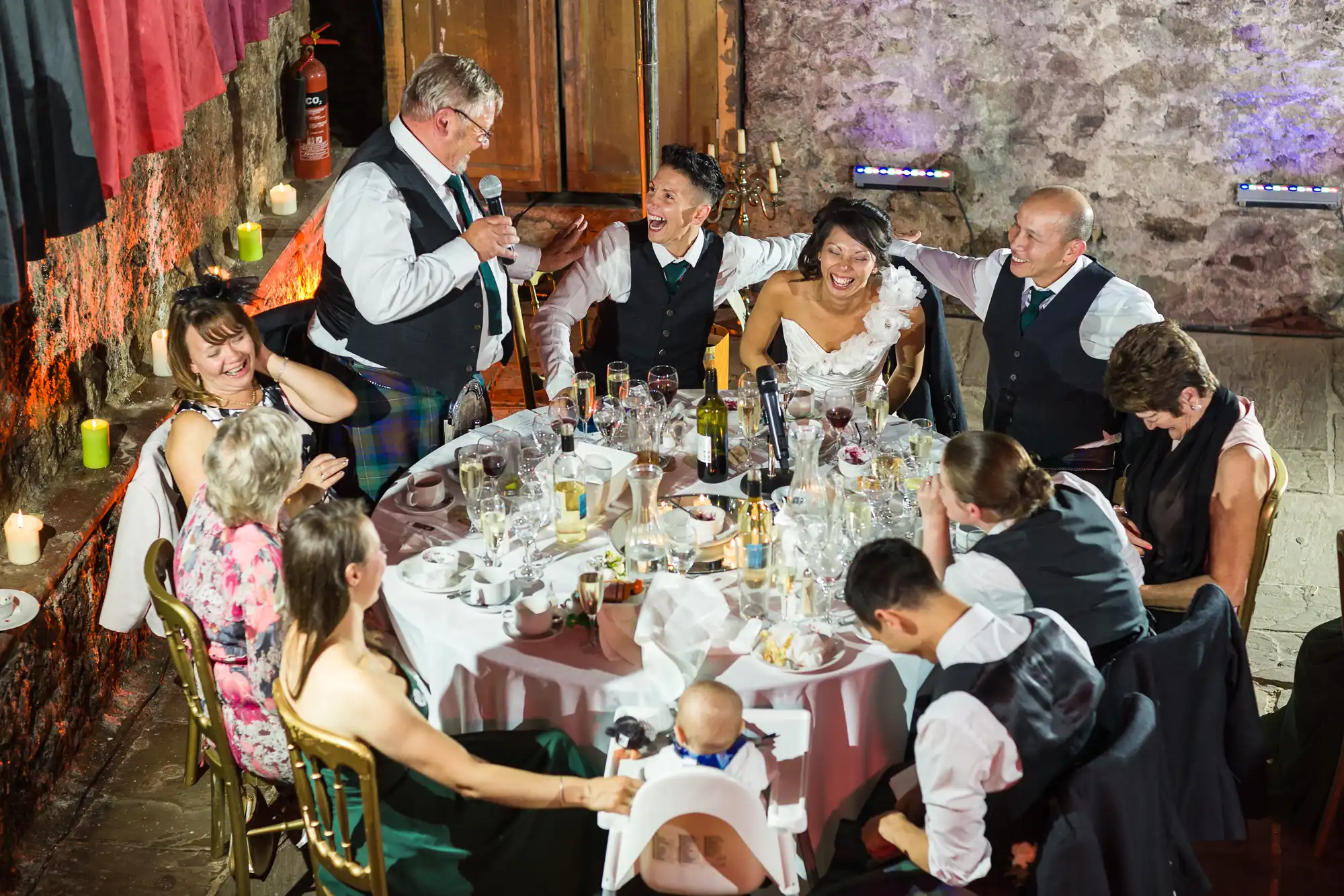 A group of people in formal attire sit around a round table at a wedding reception, laughing as an older man stands and gives a speech. The table is covered with glasses, bottles, and wedding decorations; the bride and groom are smiling joyfully among their guests.