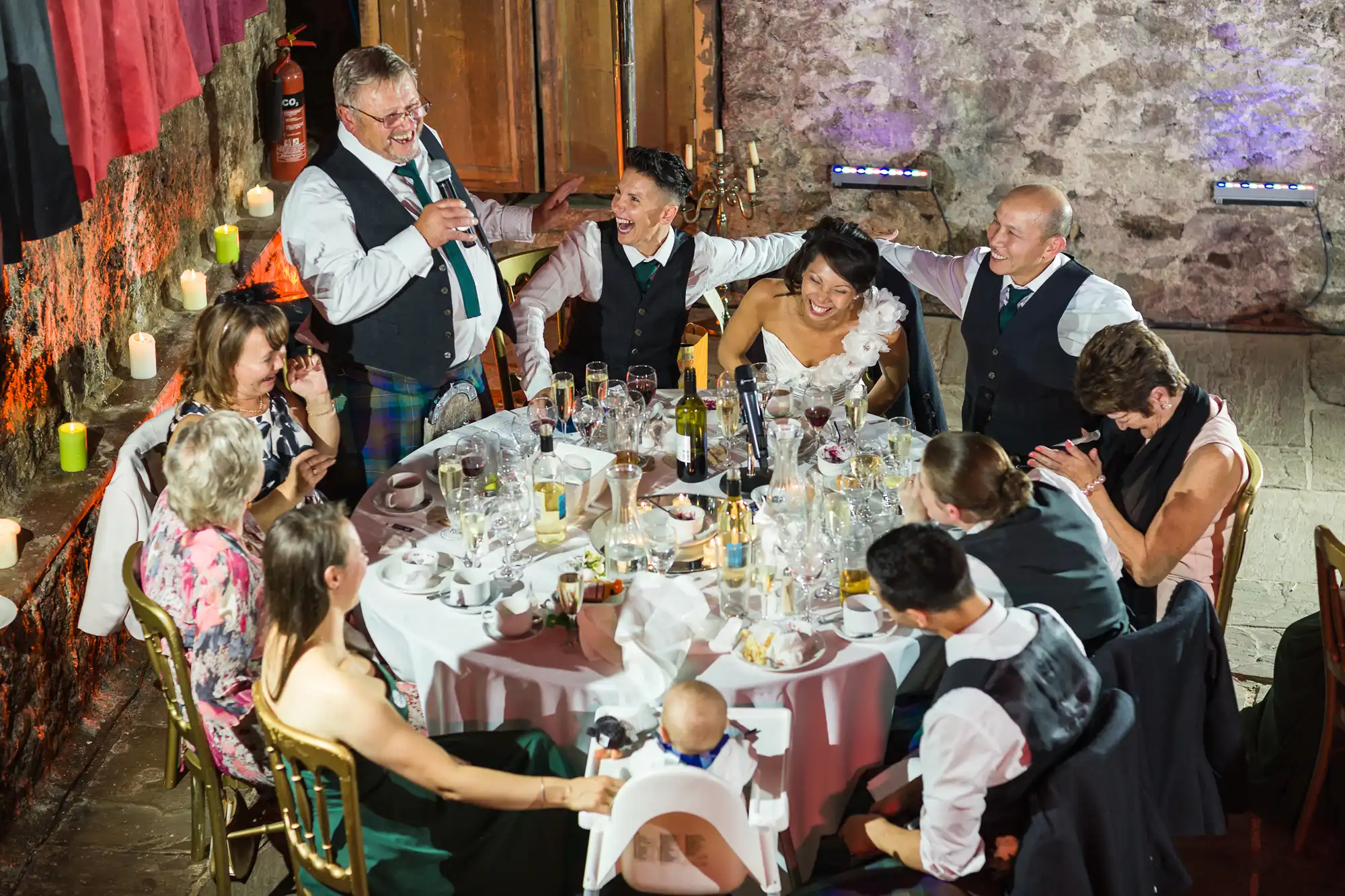 A group of people in formal attire sit around a round table covered in dishes and glasses at a wedding reception. One man stands, speaking with a microphone, while others laugh and smile. The bride in a white dress is seated, and the atmosphere is joyful and festive.