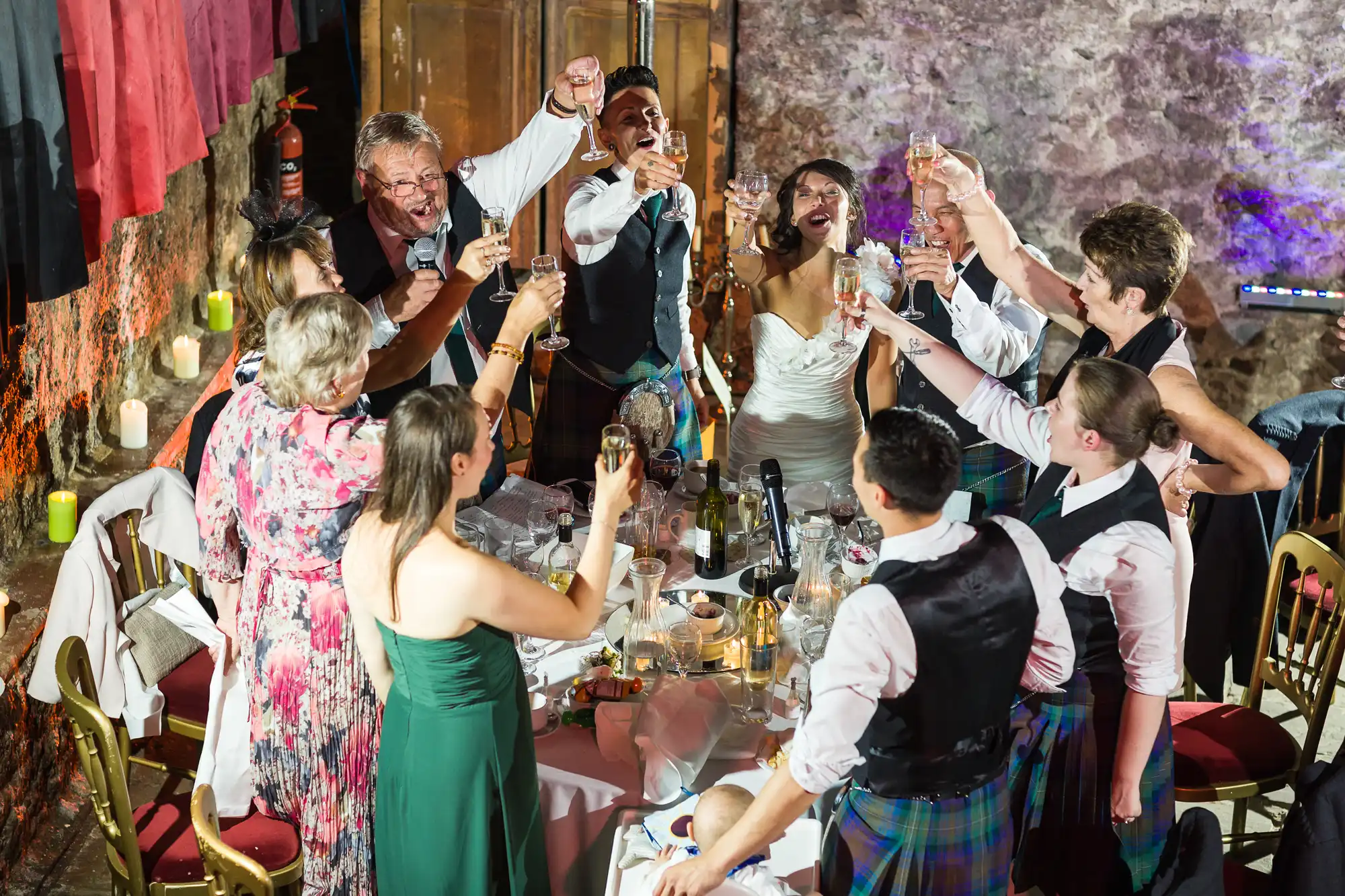 A group of people dressed in formal and semi-formal attire, including kilts and dresses, stand around a table at a wedding celebration, raising their glasses in a joyful toast. The bride and groom are smiling and the atmosphere is festive and lively.