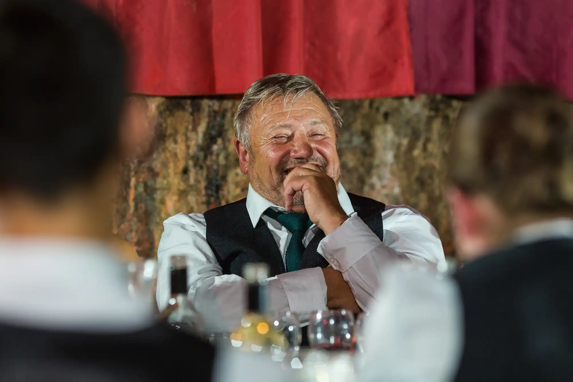 An older man in a suit and tie sits at a table, smiling with his hand over his mouth, as if laughing. He is surrounded by blurred people in formal attire, with glasses and bottles on the table in the foreground. Warm indoor lighting.