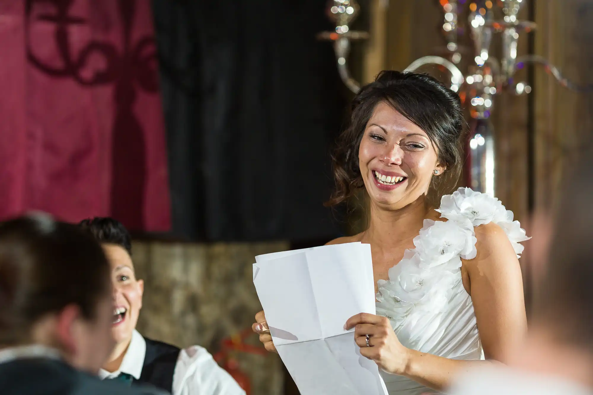 A woman in a white, one-shoulder dress smiles and laughs while holding a folded piece of paper, speaking at an indoor event. Another person in the background also smiles, and guests are visible in the foreground. The atmosphere is joyful and festive.