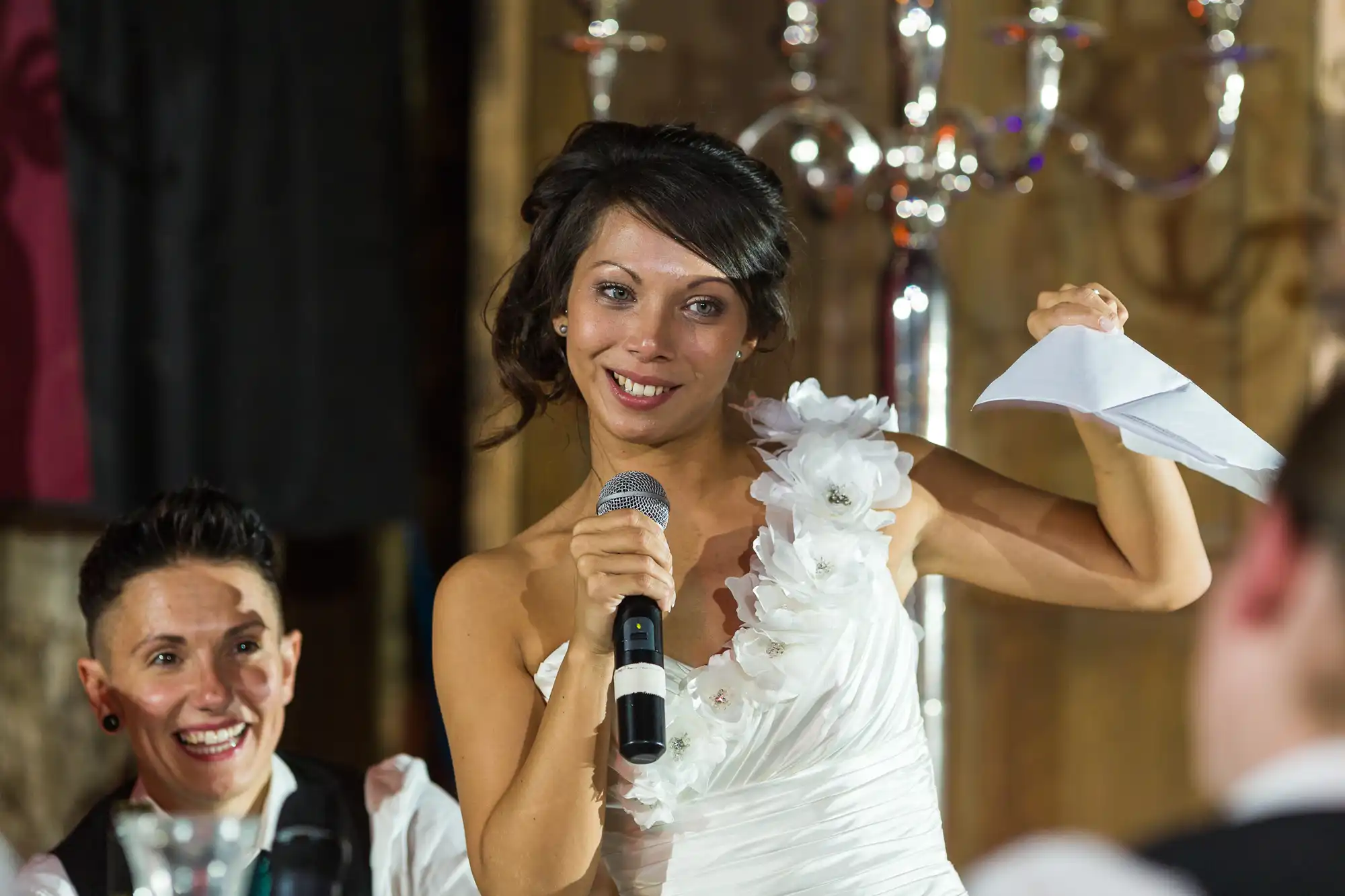 A woman in a white dress, holding a microphone and folded paper, smiles whilst giving a speech. Another person nearby smiles at her. The setting is formal, possibly a wedding, with elegant décor and warm lighting.