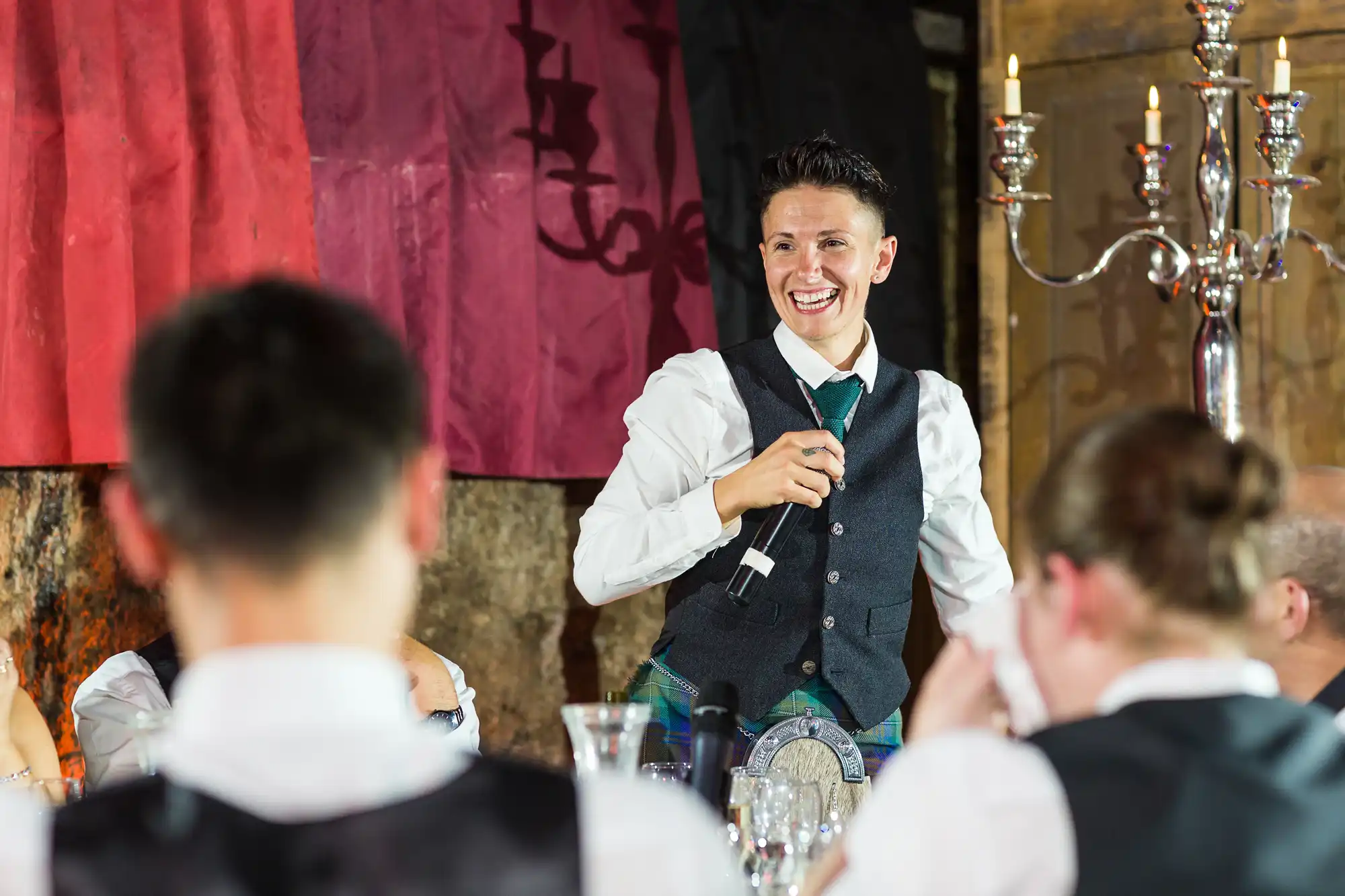A person in a waistcoat, holding a microphone, stands and smiles whilst giving a speech at a formal event. People sit at tables in the foreground, listening, with elegant decorations and candelabra visible in the warmly lit room.