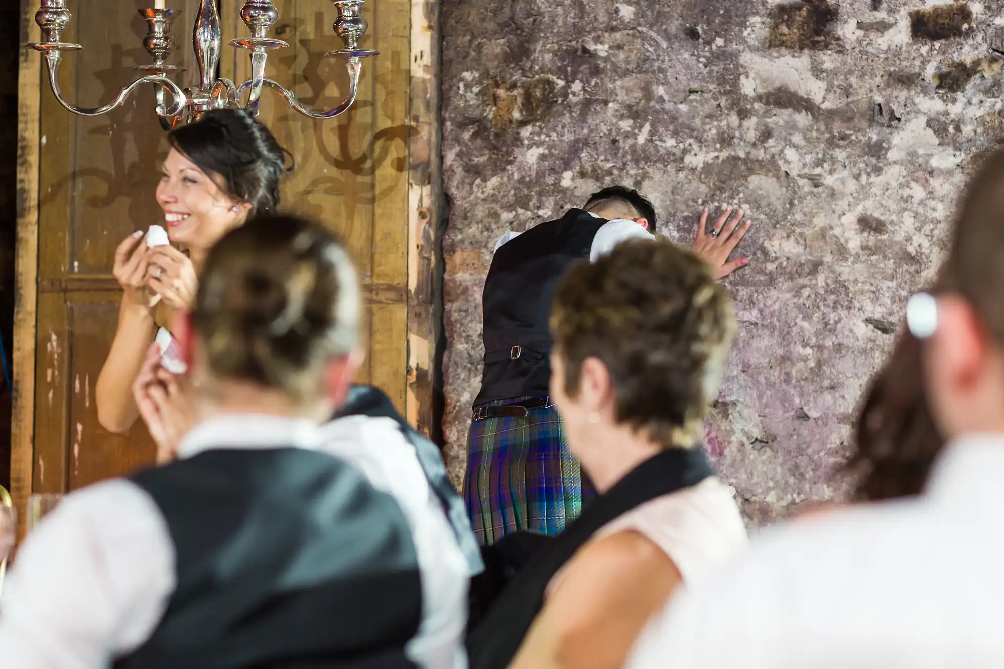 At a formal event, a smiling woman in the foreground holds a microphone. In the background, a man in a waistcoat and tartan kilt faces a stone wall, covering his face with one hand. Several other people in formal attire are seated, watching the scene.