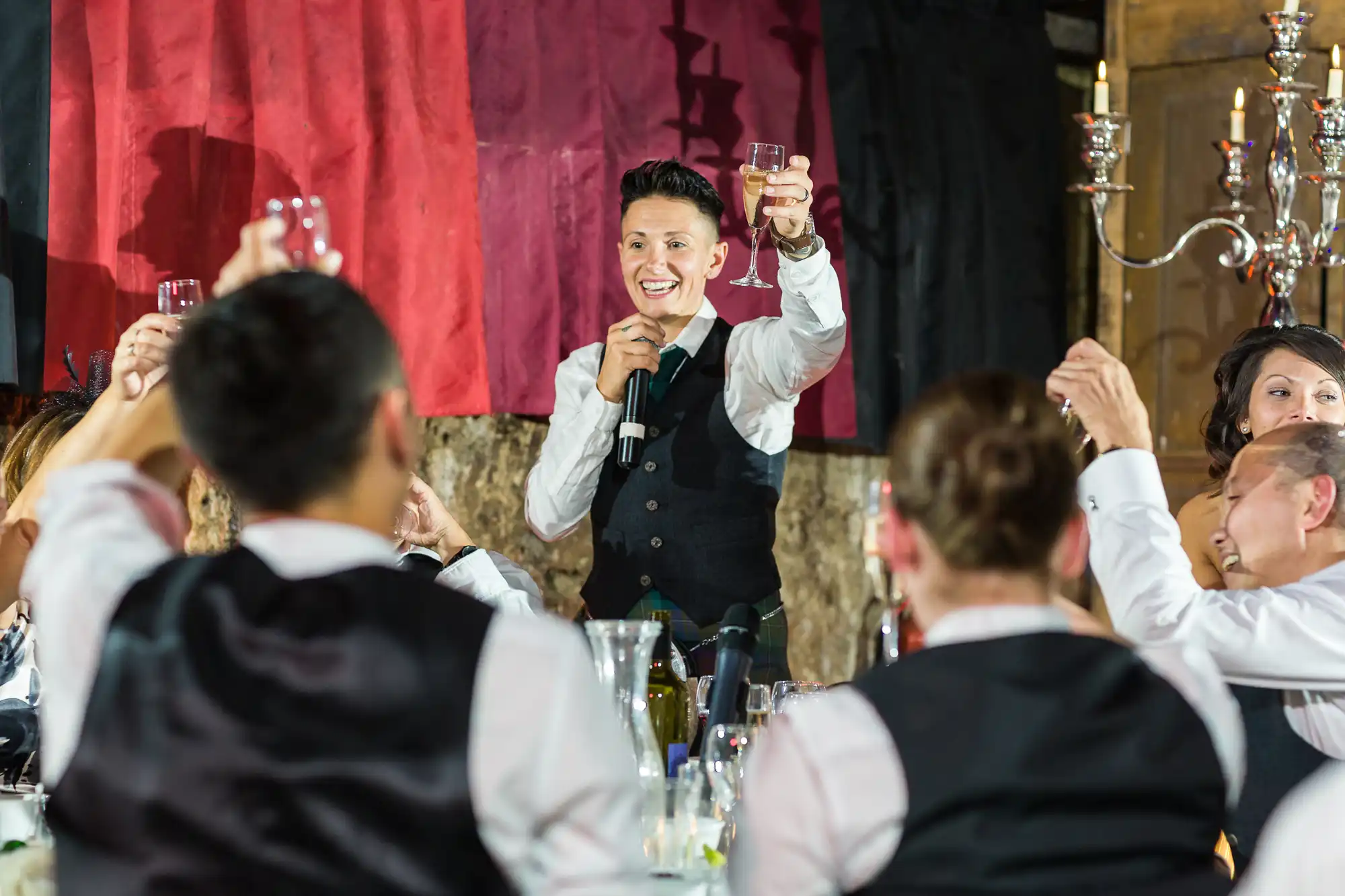 A person in a black waistcoat and tie stands holding a microphone and raising a glass in a toast, smiling at seated guests who also lift their glasses. The setting appears festive, with candles, drinks, and a red and black cloth backdrop.