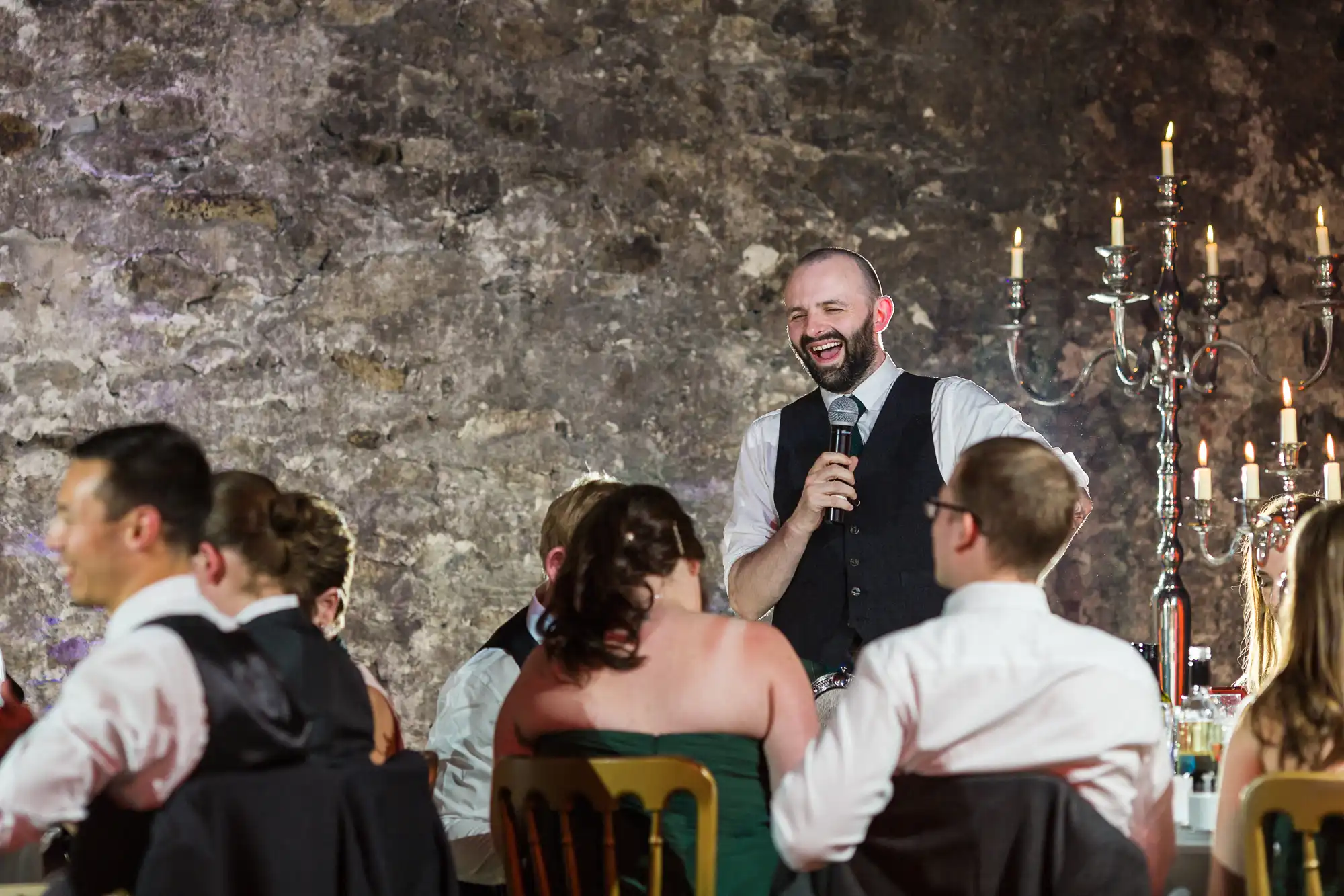 A man in a waistcoat stands laughing and holding a microphone, speaking to seated guests at a formal event. The group listens attentively around decorated tables, with an ornate candelabrum and a stone wall in the background, suggesting a celebration or wedding.