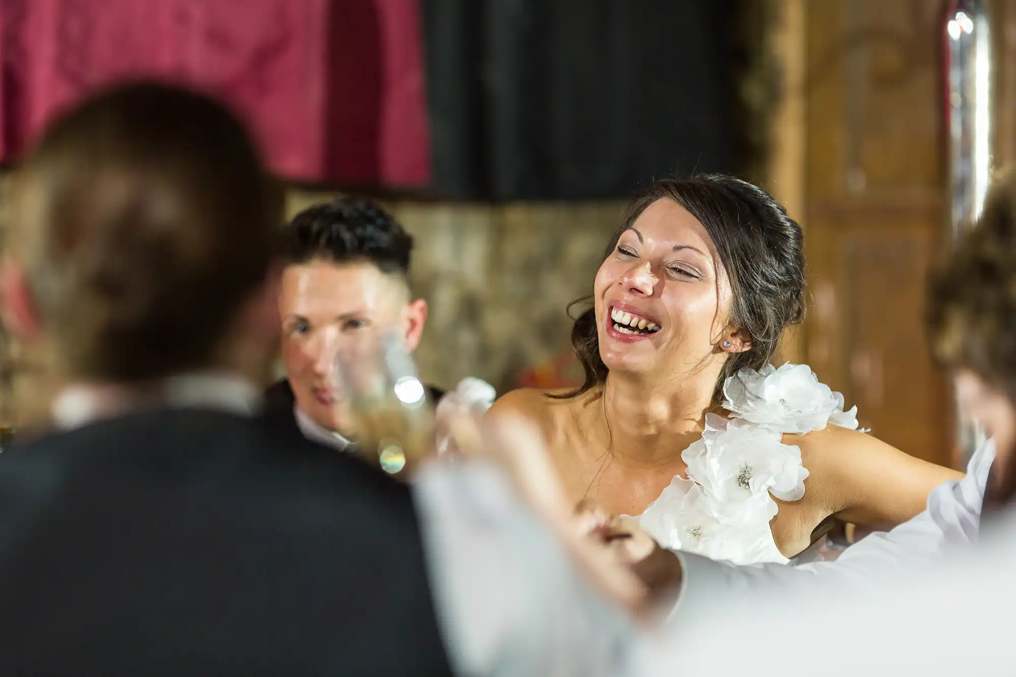 A woman in a white dress with flower details laughs joyfully at an indoor event, surrounded by people in formal attire. The atmosphere appears festive, with blurred guests in the foreground and warm lighting highlighting her expression.