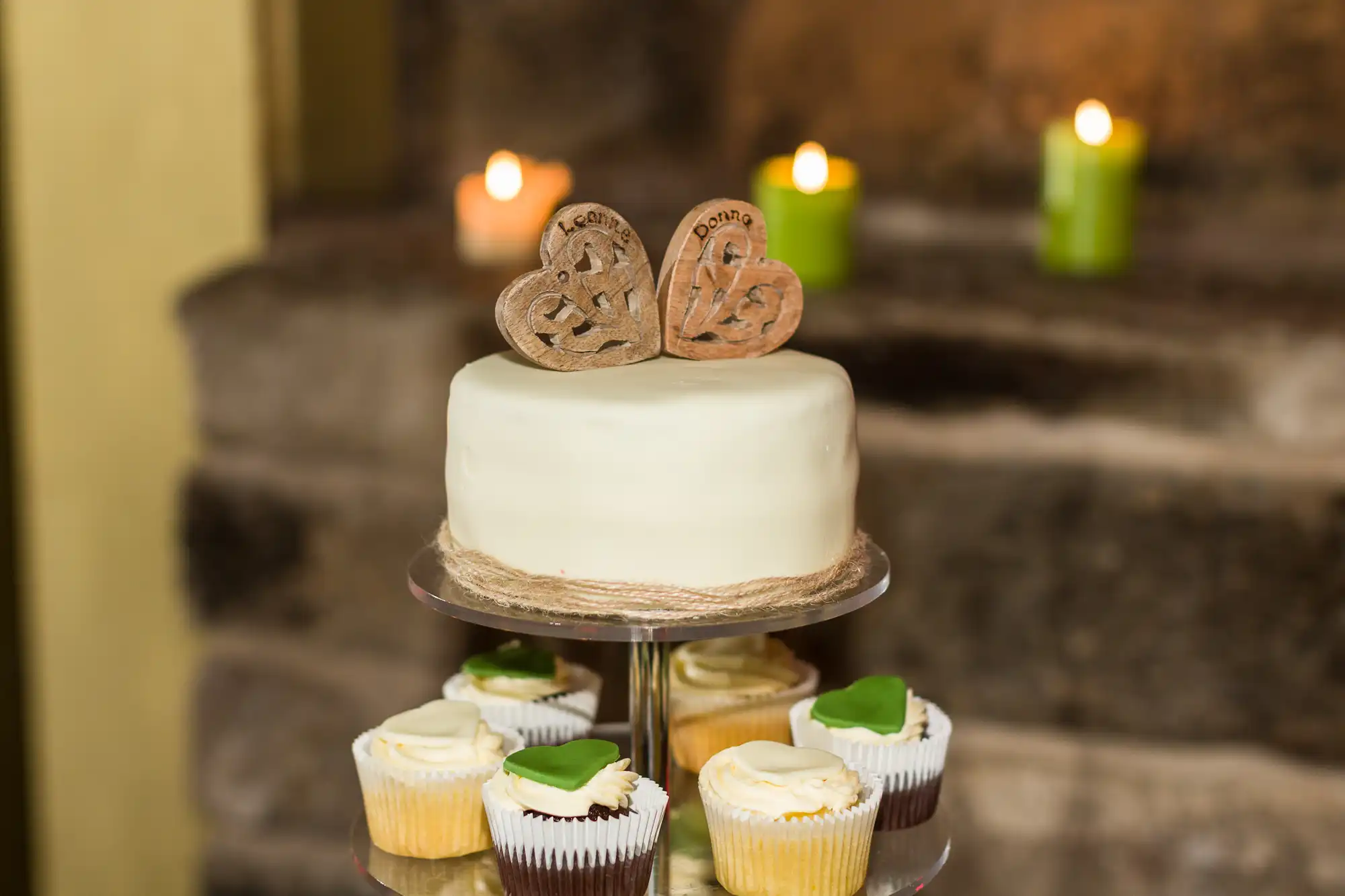 A white iced cake topped with two carved wooden hearts sits on a stand wrapped with string. Surrounding it are cupcakes with white icing and green fondant leaves. Three lit candles and stone steps are blurred in the background.