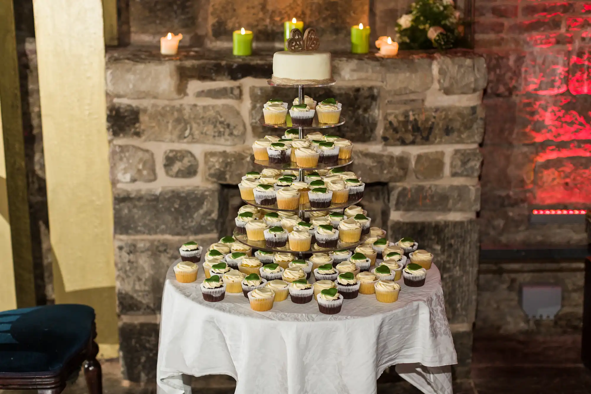 A tiered cupcake stand holds assorted cupcakes with white icing and green garnish, topped with a small white cake and silver “50” topper. The display sits on a white tablecloth in front of a rustic stone wall with candles and green decorations.