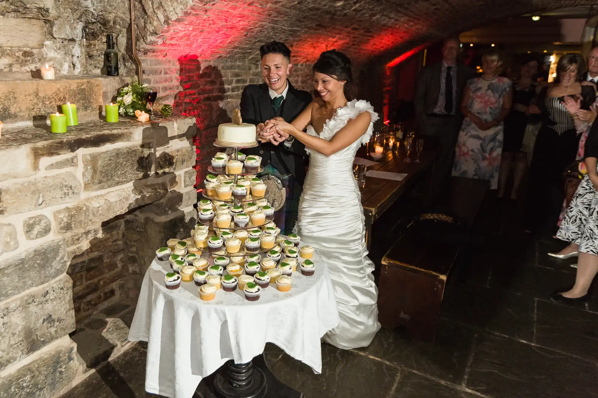 A bride and groom smile while cutting a small cake atop a tiered stand of cupcakes in a rustic, candlelit brick venue. The bride wears a white dress with floral details; the groom wears a dark suit. Guests watch and smile in the background.