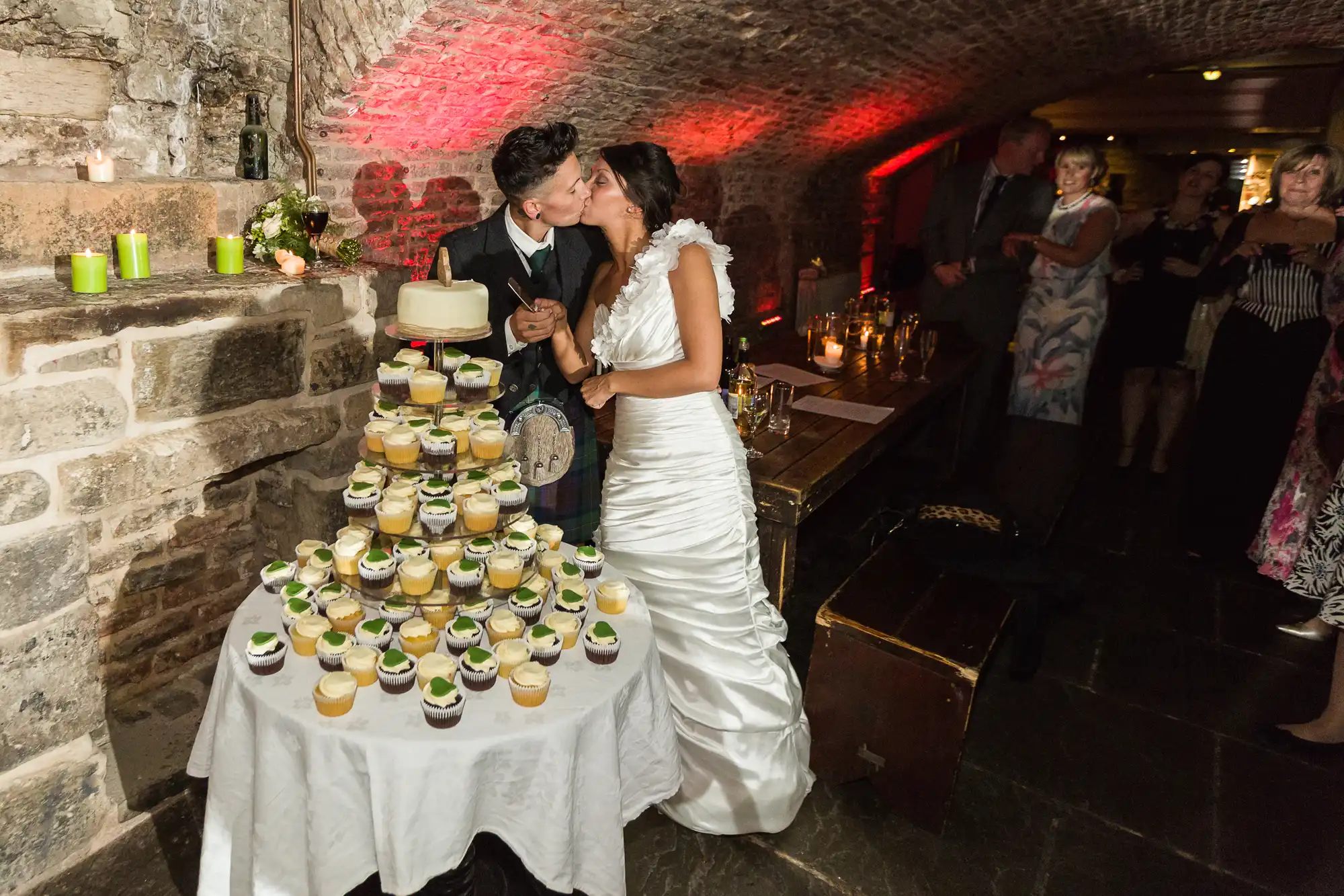 A newly-wed couple in wedding attire kiss beside a tiered cupcake stand topped with a small cake, set in a rustic stone-walled room with arched ceilings. Guests watch in the background; soft candlelight and red uplighting add warmth to the scene.
