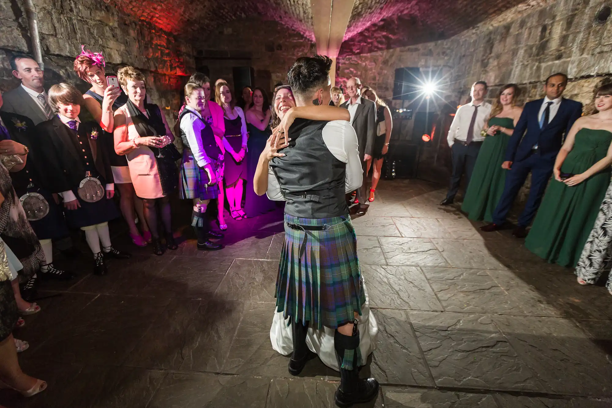 A couple dances in the centre of a stone-floored room with arched brick ceilings, surrounded by formally dressed guests. The groom wears a kilt and waistcoat. People stand in a circle, smiling and watching, as a bright flash illuminates the scene from behind.