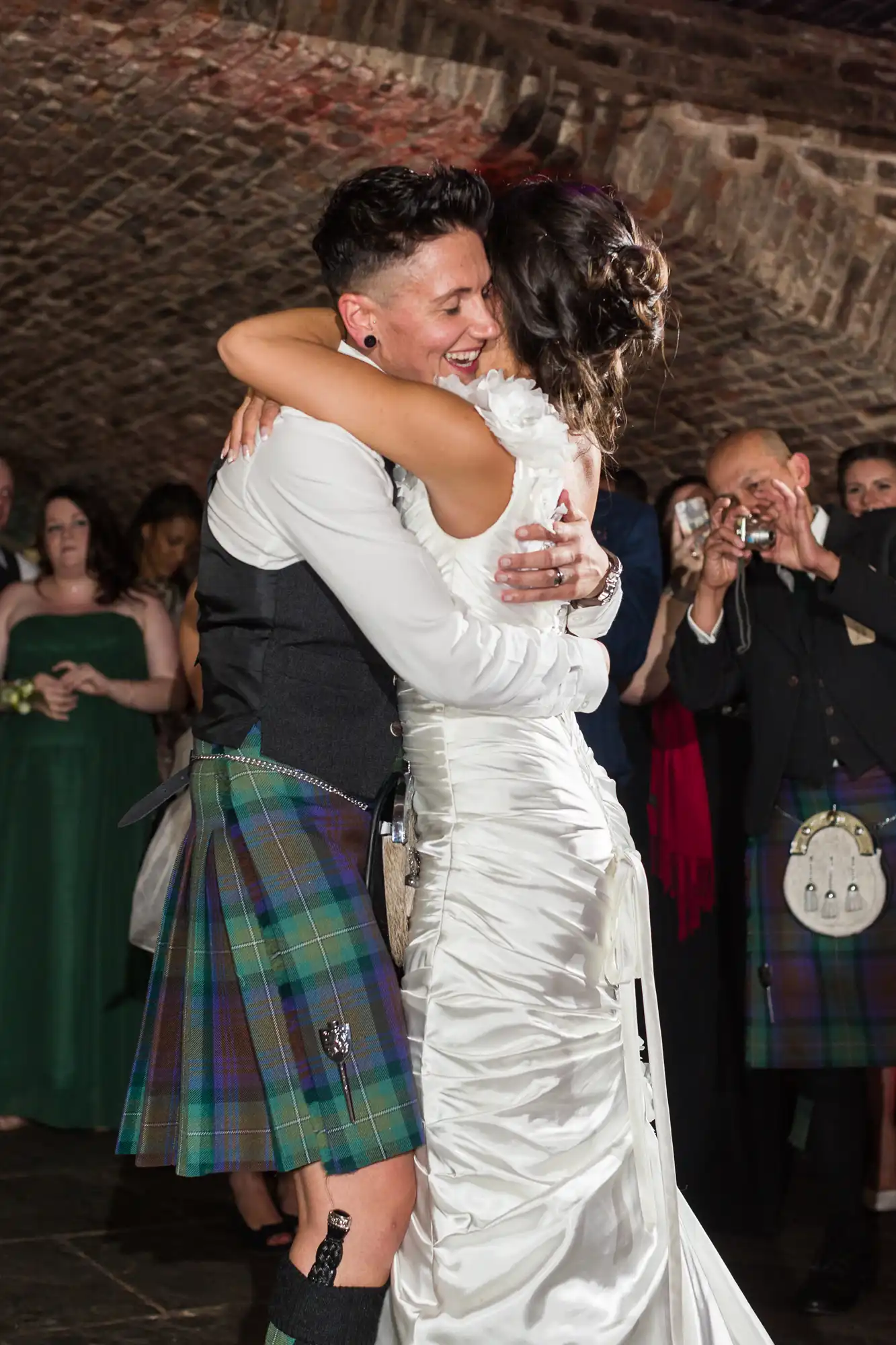 A joyful couple, one in a white satin wedding dress and the other in a black waistcoat with a tartan kilt, embrace and smile broadly whilst dancing. Onlookers in formal attire, some taking photos, watch under a vaulted brick ceiling.