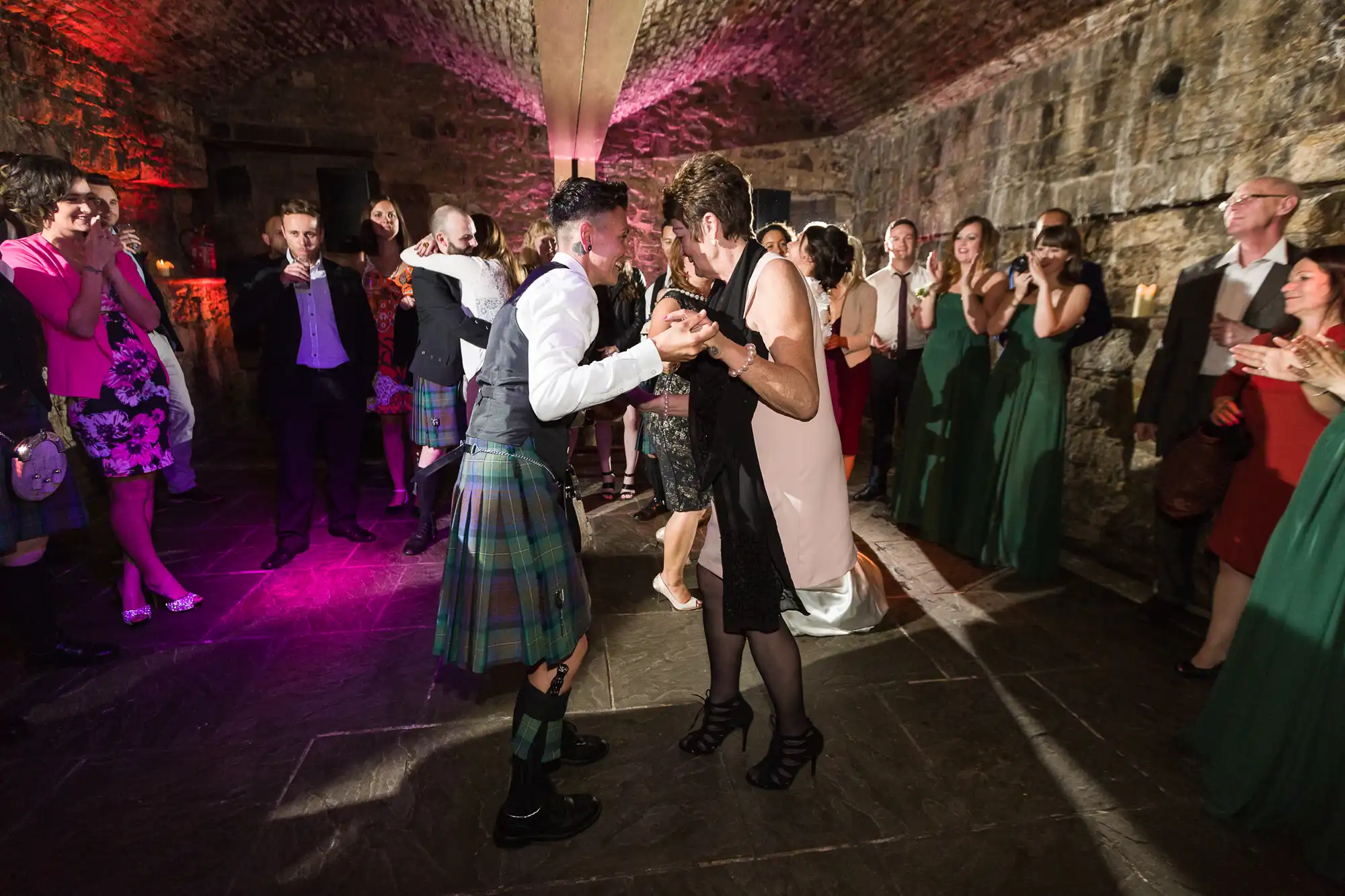 A lively wedding dance scene: a couple, one in a kilt and one in a dress, dance together in a stone-walled, arched room, surrounded by smiling and clapping guests dressed in formal and semi-formal attire. Warm, colourful lighting enhances the festive atmosphere.