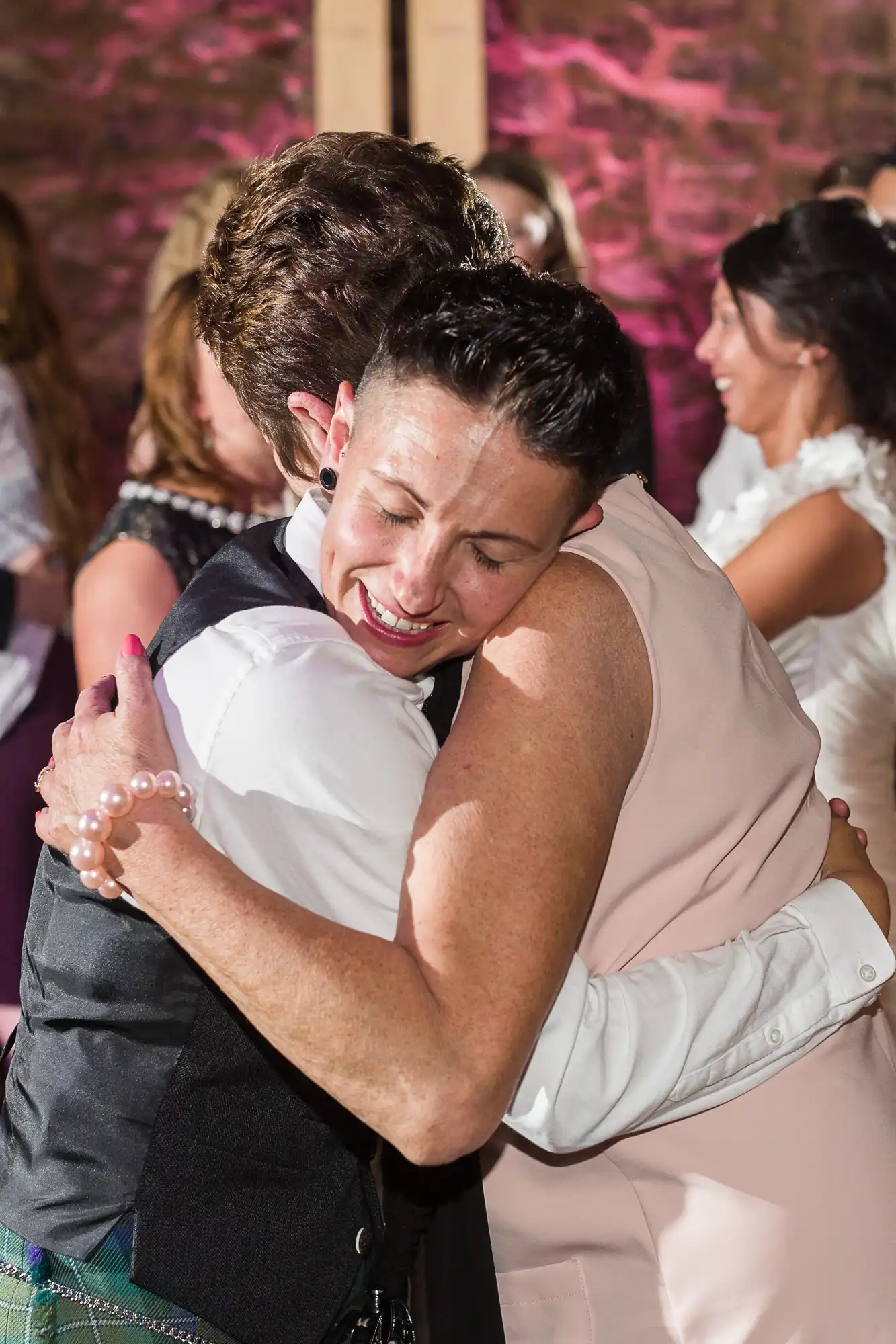 Two people warmly embrace and smile with their eyes closed at a joyful event. Both have short hair; one wears a white shirt and waistcoat, the other a light pink dress and a pearl bracelet. Other guests, blurred in the background, are also smiling.